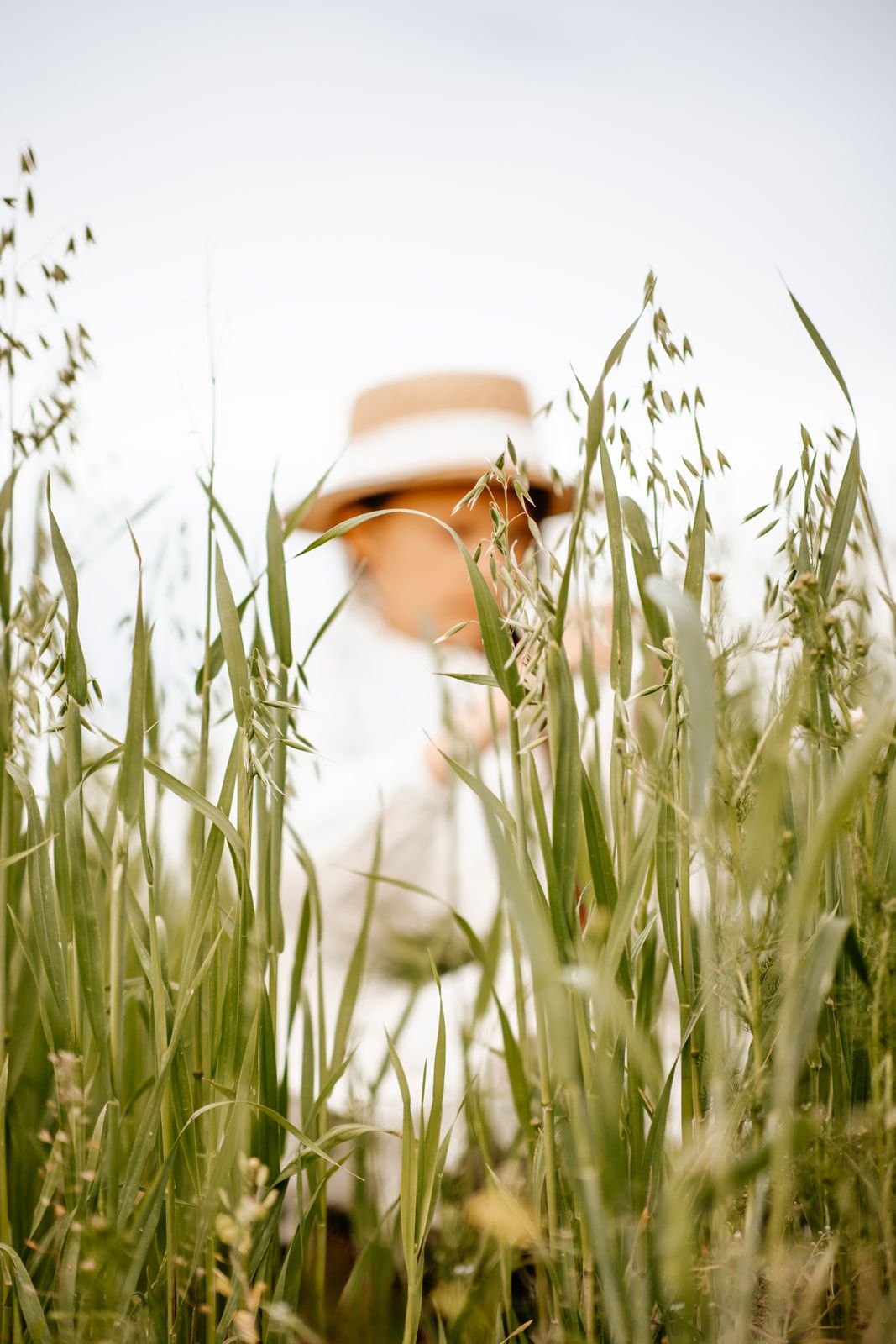 Blurred figure of woman in white dress and straw hat standing among tall green grass in a field.