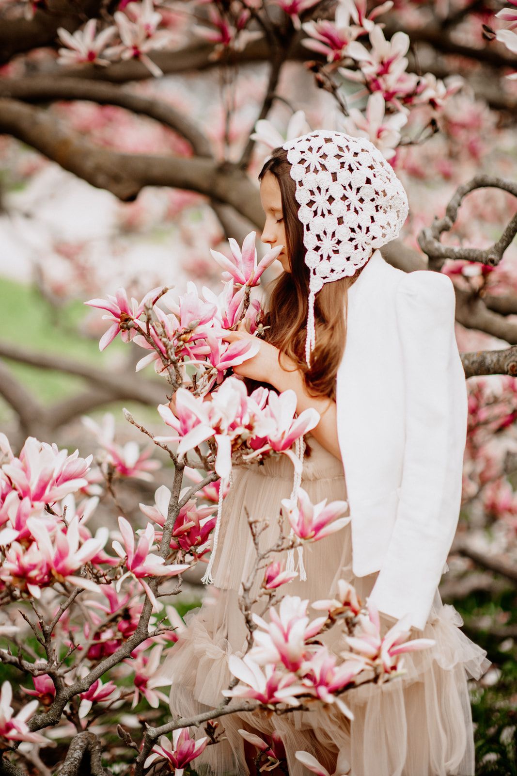 Young girl in white lace bonnet and dress smelling pink magnolia blossoms on a blooming tree in spring.