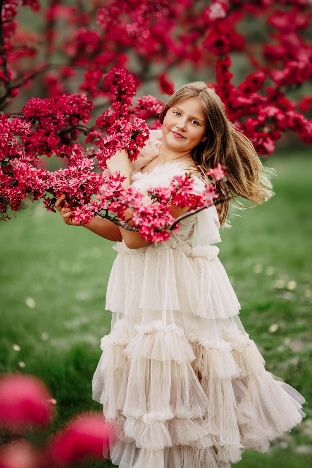 Young girl in white ruffled dress holding pink blossoms beside a flowering red tree in a garden.