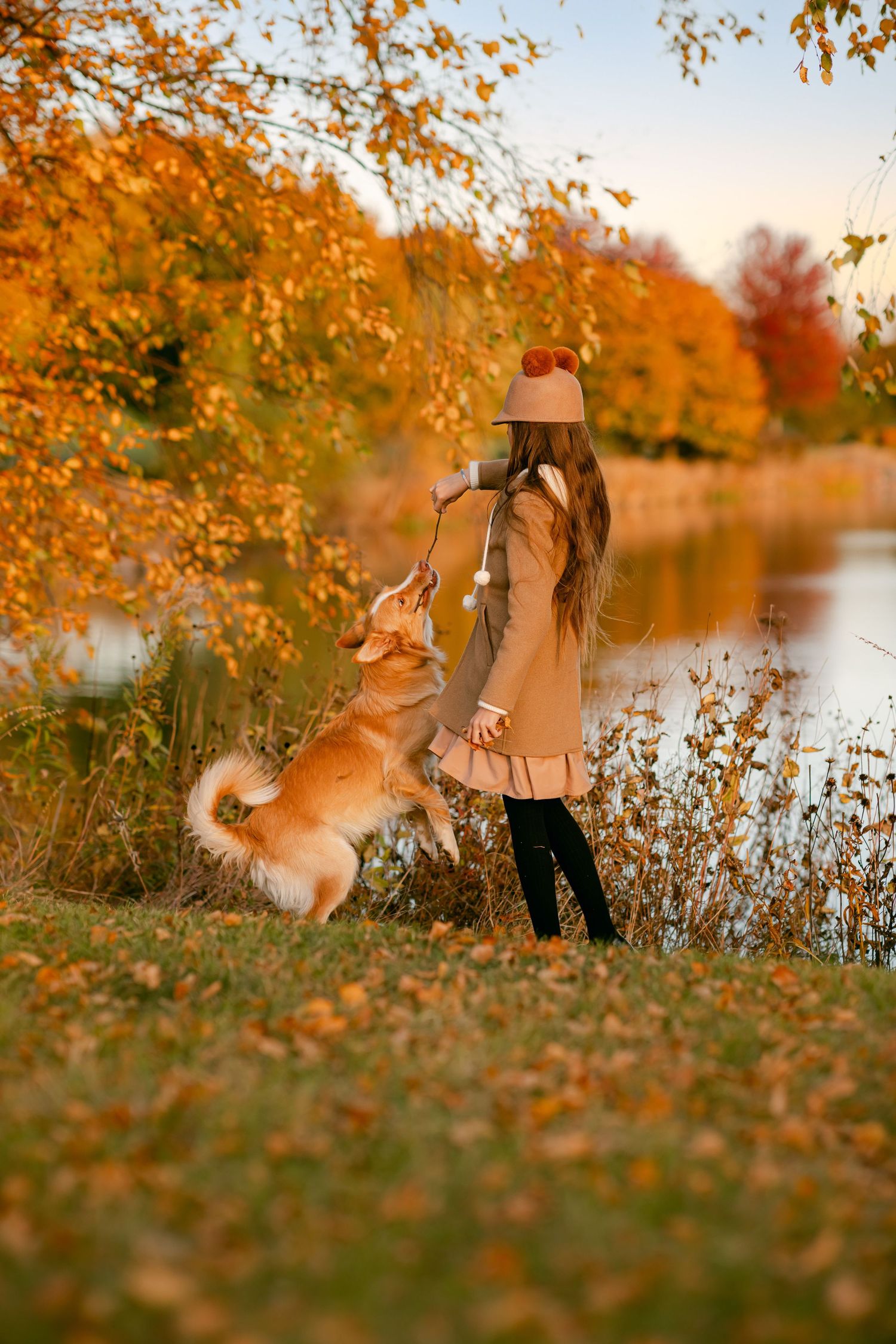 Girl in beige coat plays with golden dog by autumn lake, surrounded by vibrant orange fall foliage.