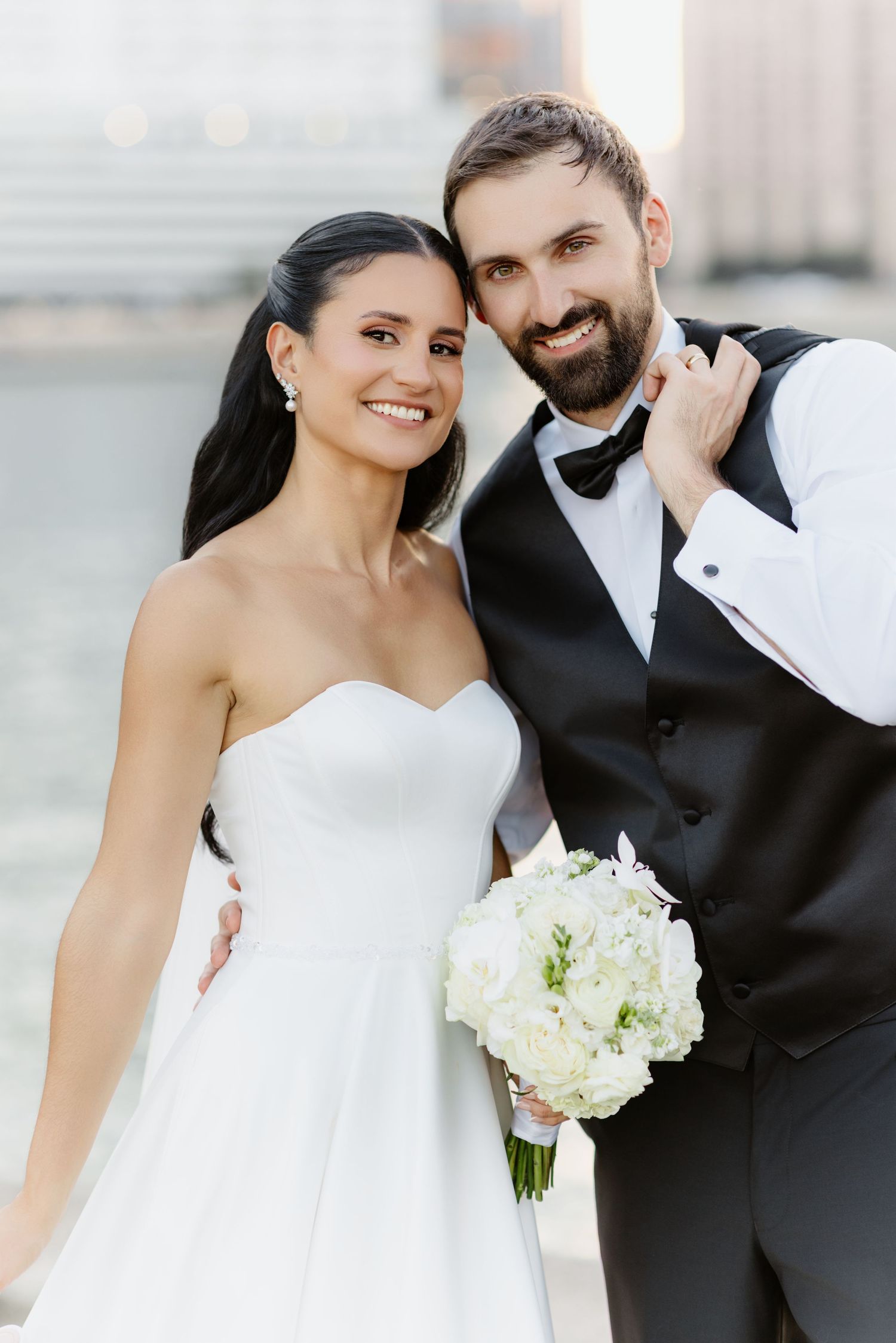 Smiling bride and bearded groom pose outdoors, she in strapless gown, he in tuxedo holding white bouquet.