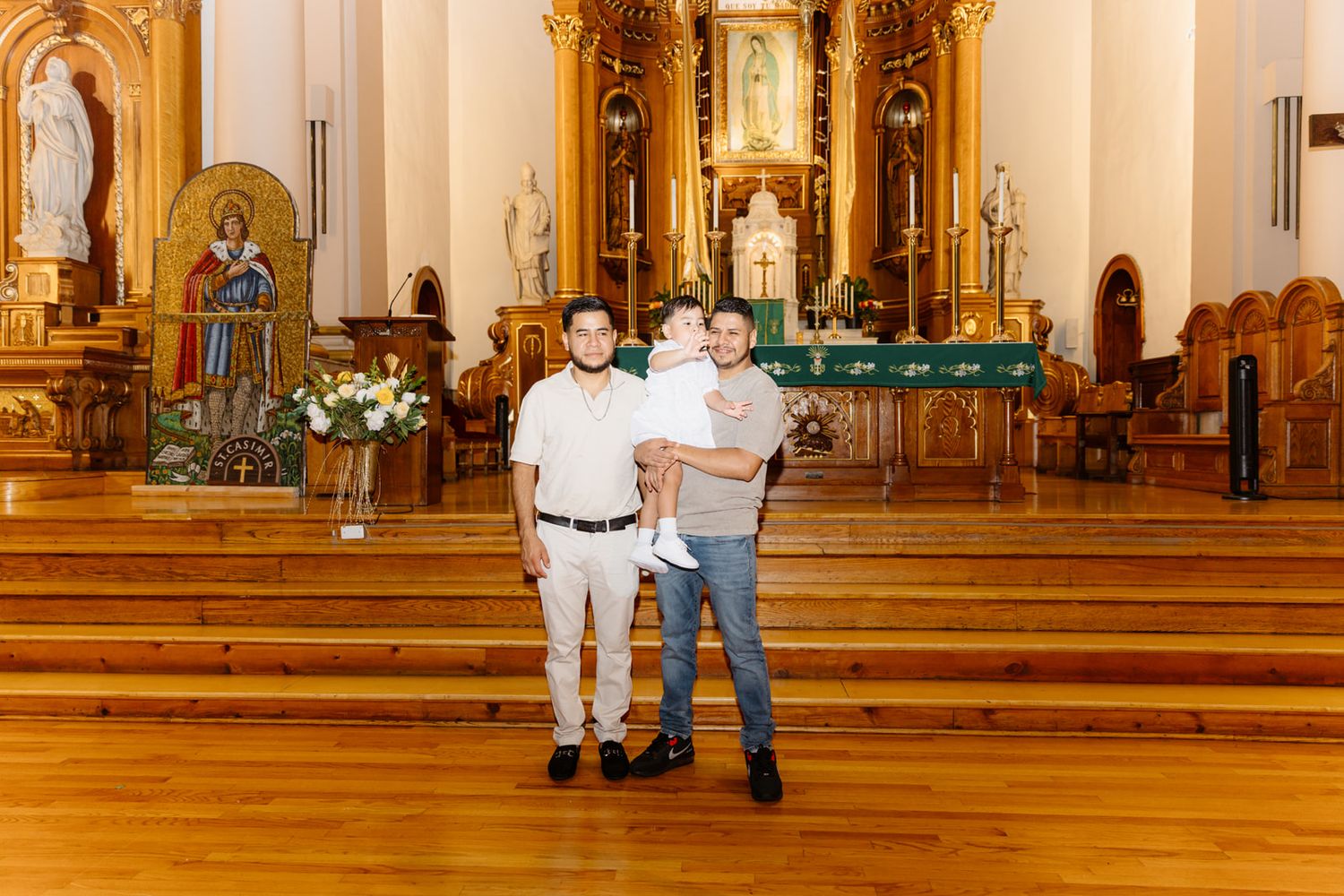 Two men holding a baby pose inside an ornate Catholic church with wooden floors and golden altar.