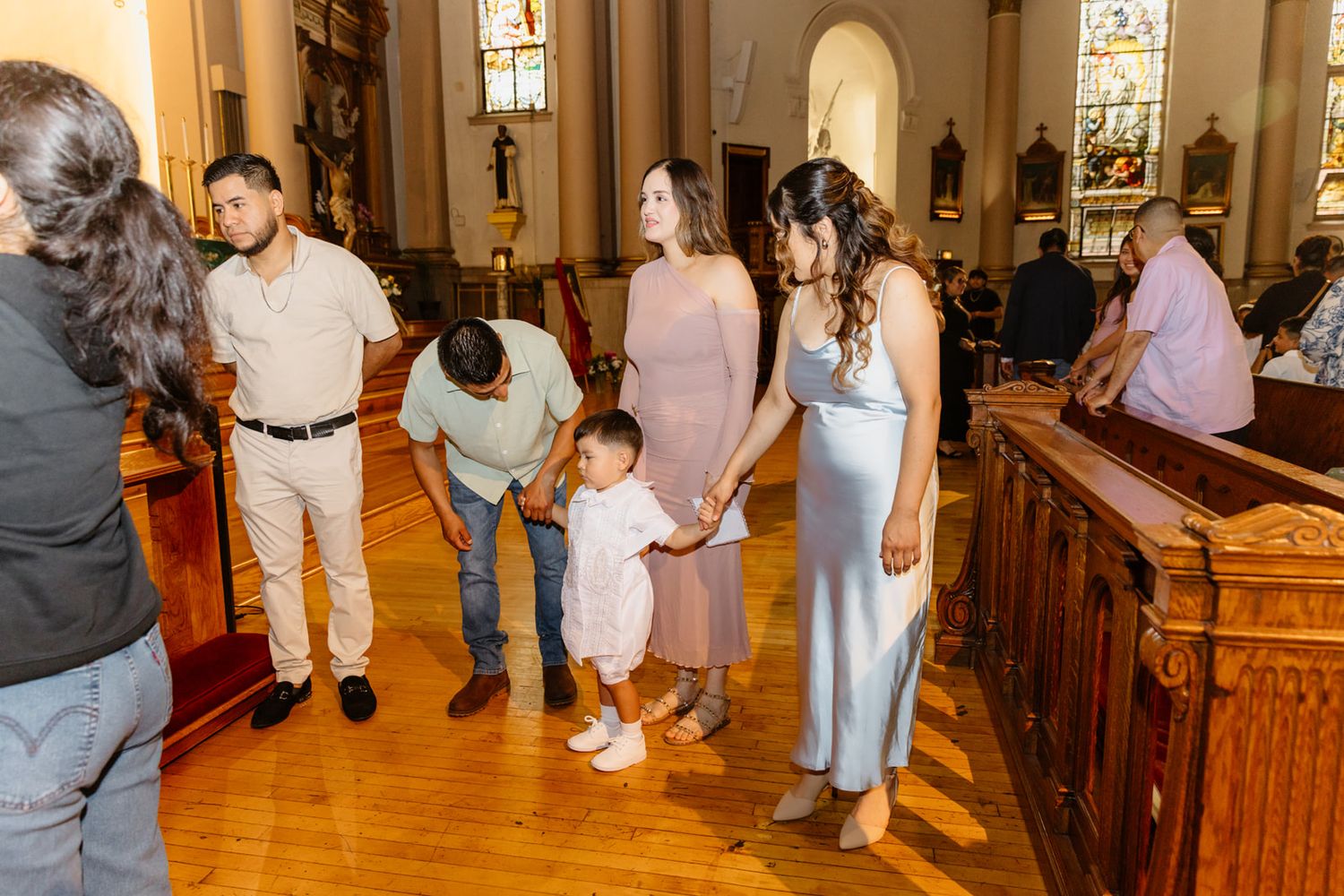 Family gathering in a church aisle, with adults and a toddler in formal attire during a religious ceremony.