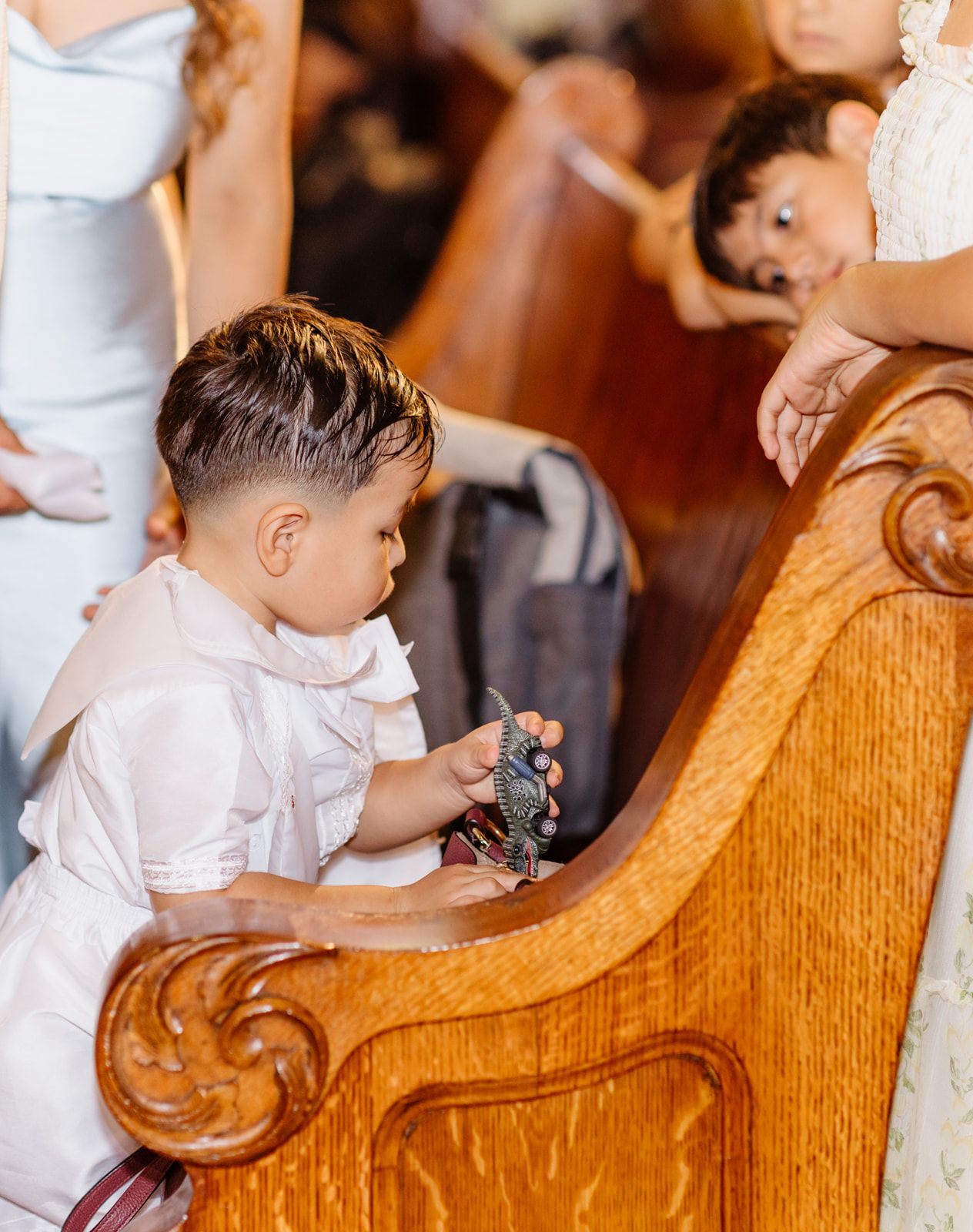 Young boy in white outfit sits at church pew during a wedding ceremony holding small decorative item.
