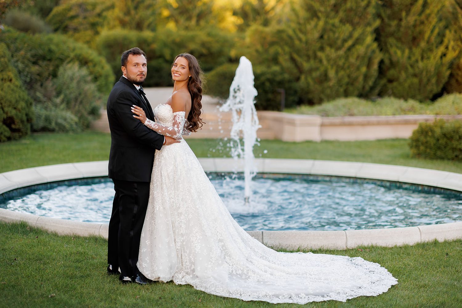Couple shares tender embrace before garden fountain, bride in elegant lace wedding dress with train.