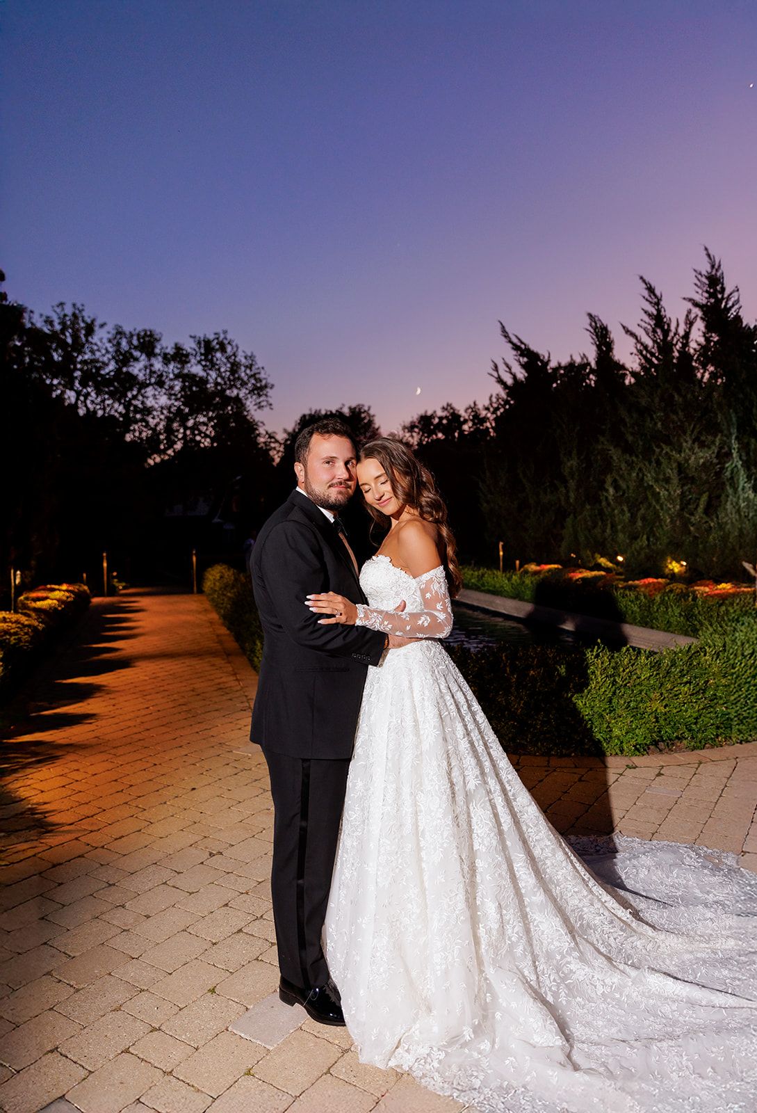 Groom embraces bride from behind on a garden path at dusk, her lace gown trailing elegantly.