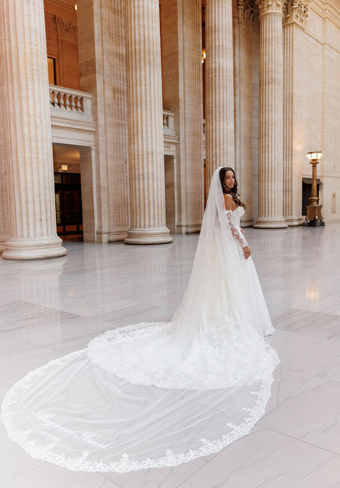 Bride in long-sleeve lace gown with cathedral veil poses among grand marble columns in ornate hall.