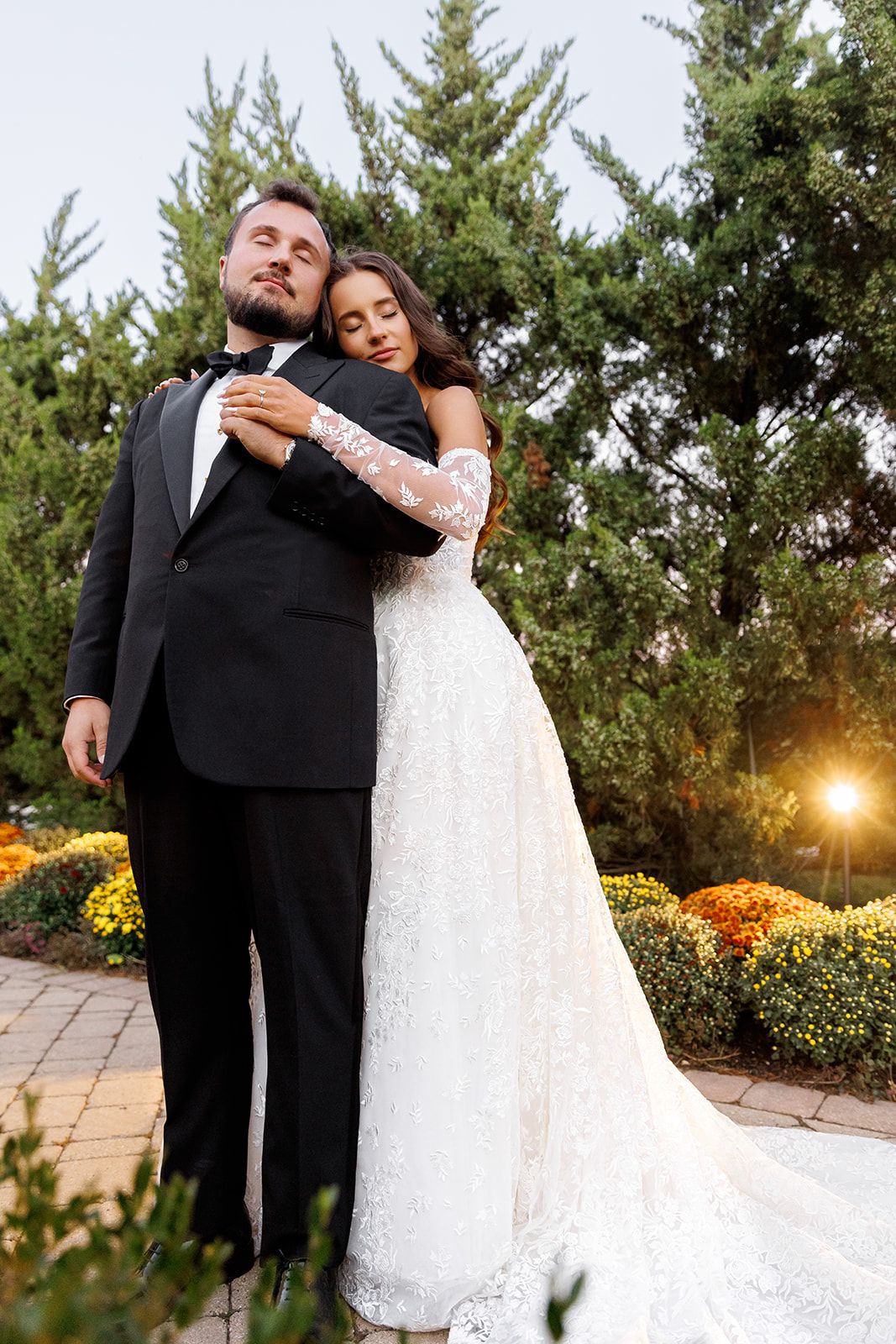 Bride in lace gown embraces groom in black tuxedo outdoors, surrounded by evergreen trees and colorful flowers.
