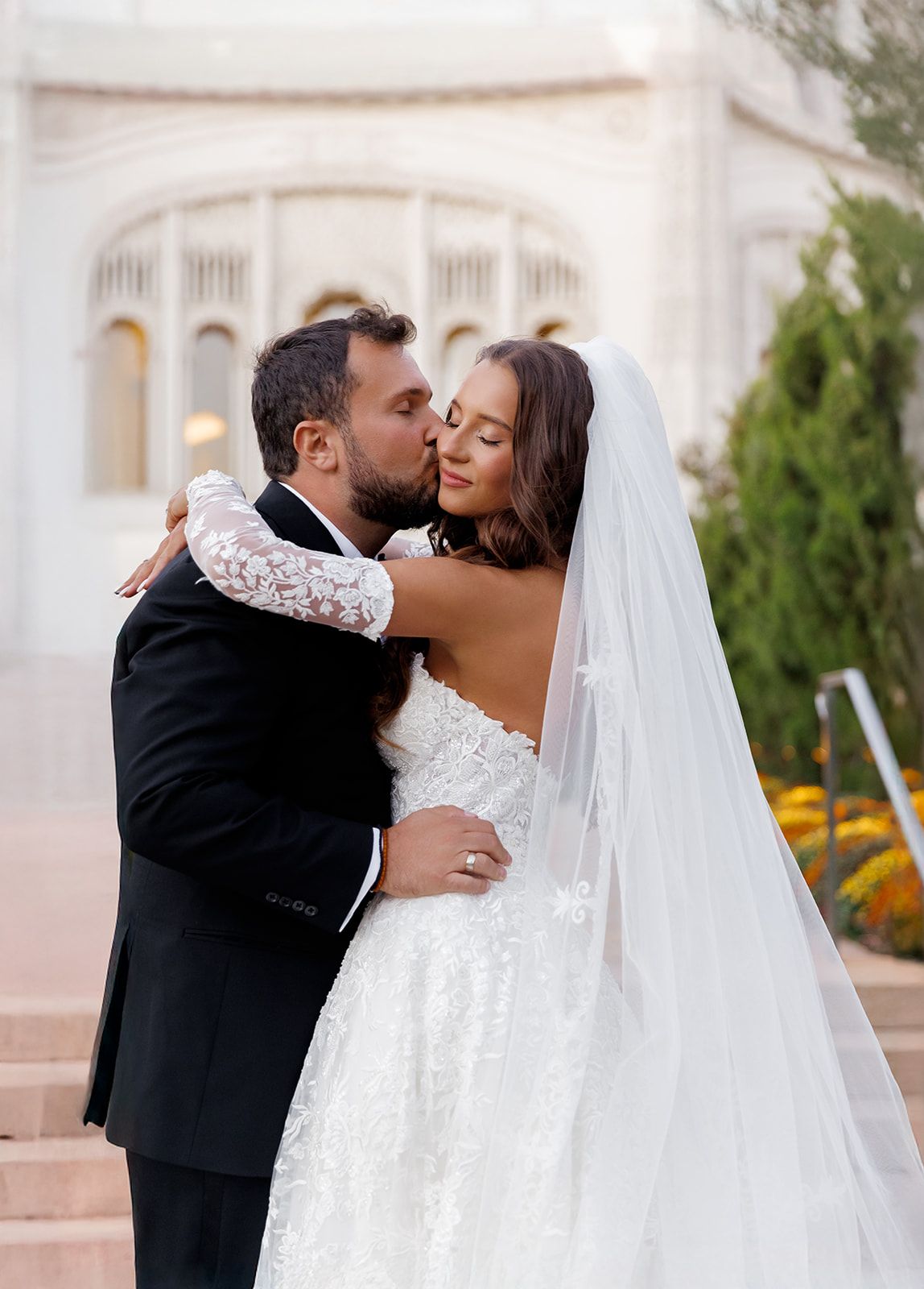 Groom kisses smiling bride on the cheek on steps before a white ornate building, bride in backless lace gown with veil.