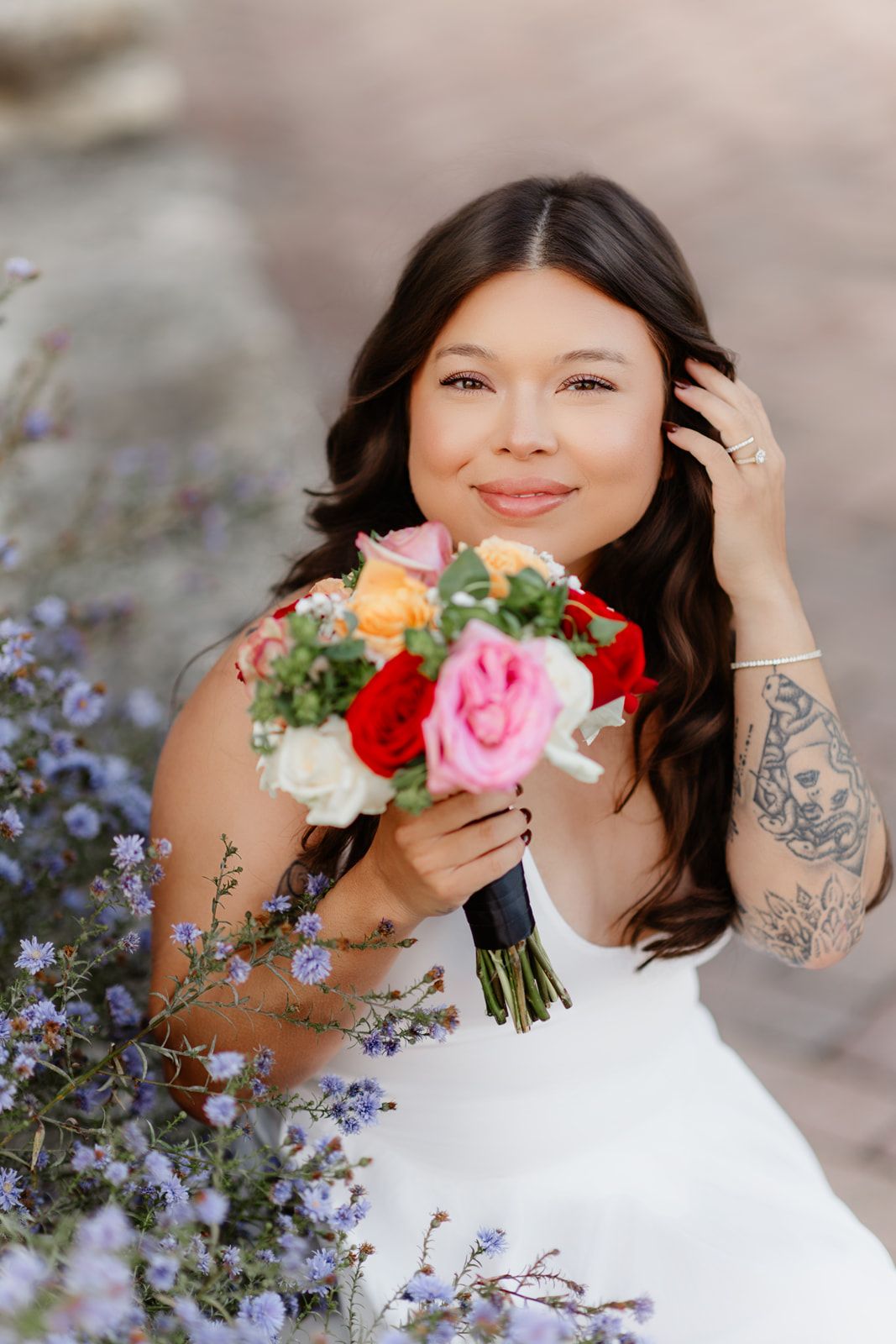 Smiling bride in white dress holding colorful rose bouquet, with tattoo on arm, surrounded by purple wildflowers.