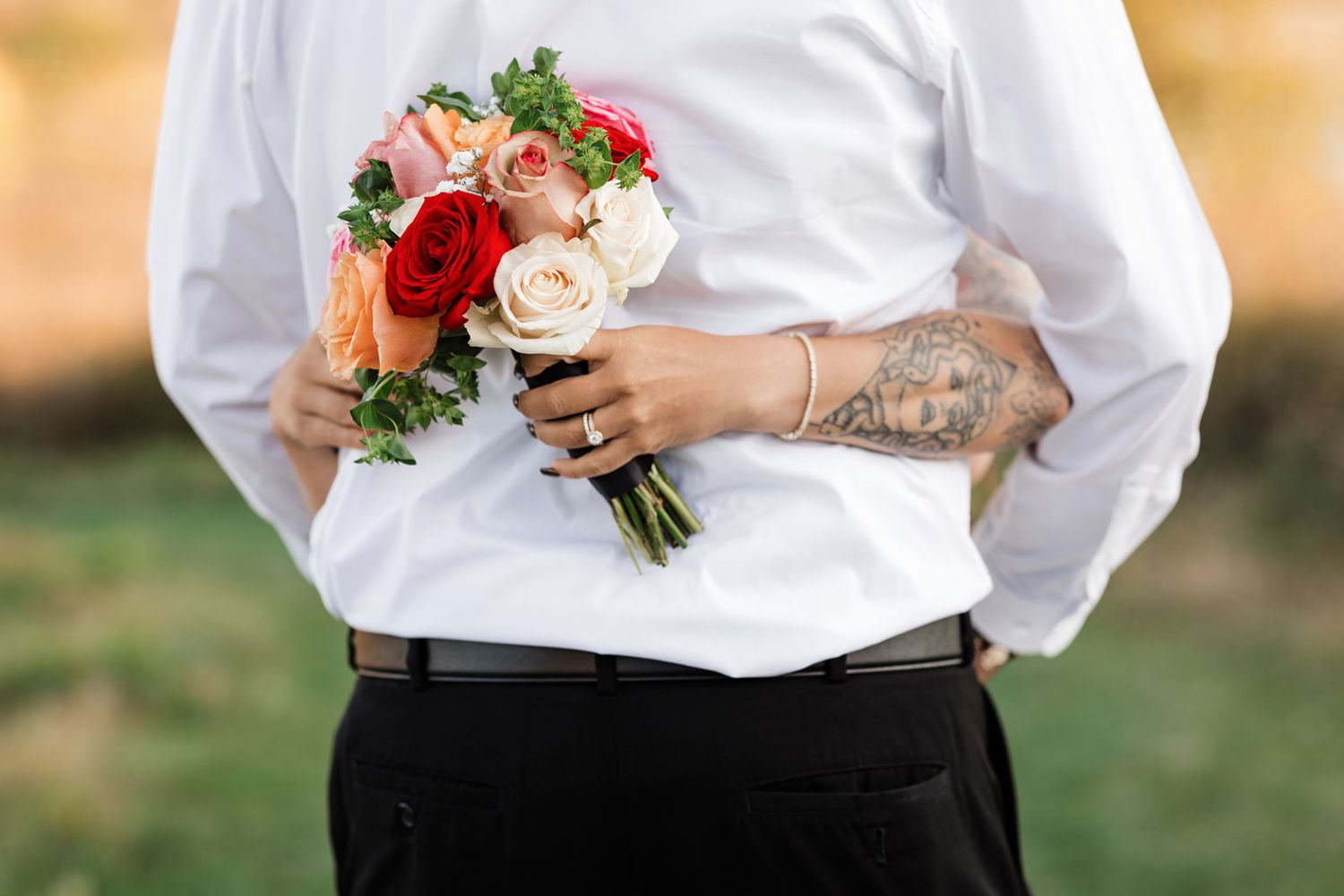 Person in white shirt holding a colorful bridal bouquet of red, pink, and cream roses behind their back.