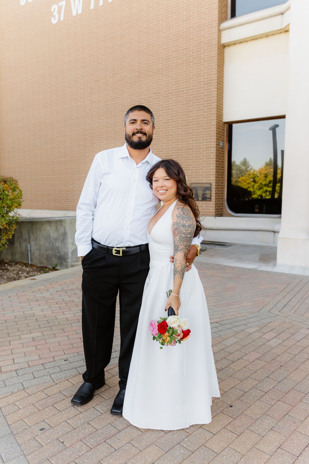 Couple in wedding attire smiling outside brick building, bride holding colorful bouquet in white dress.