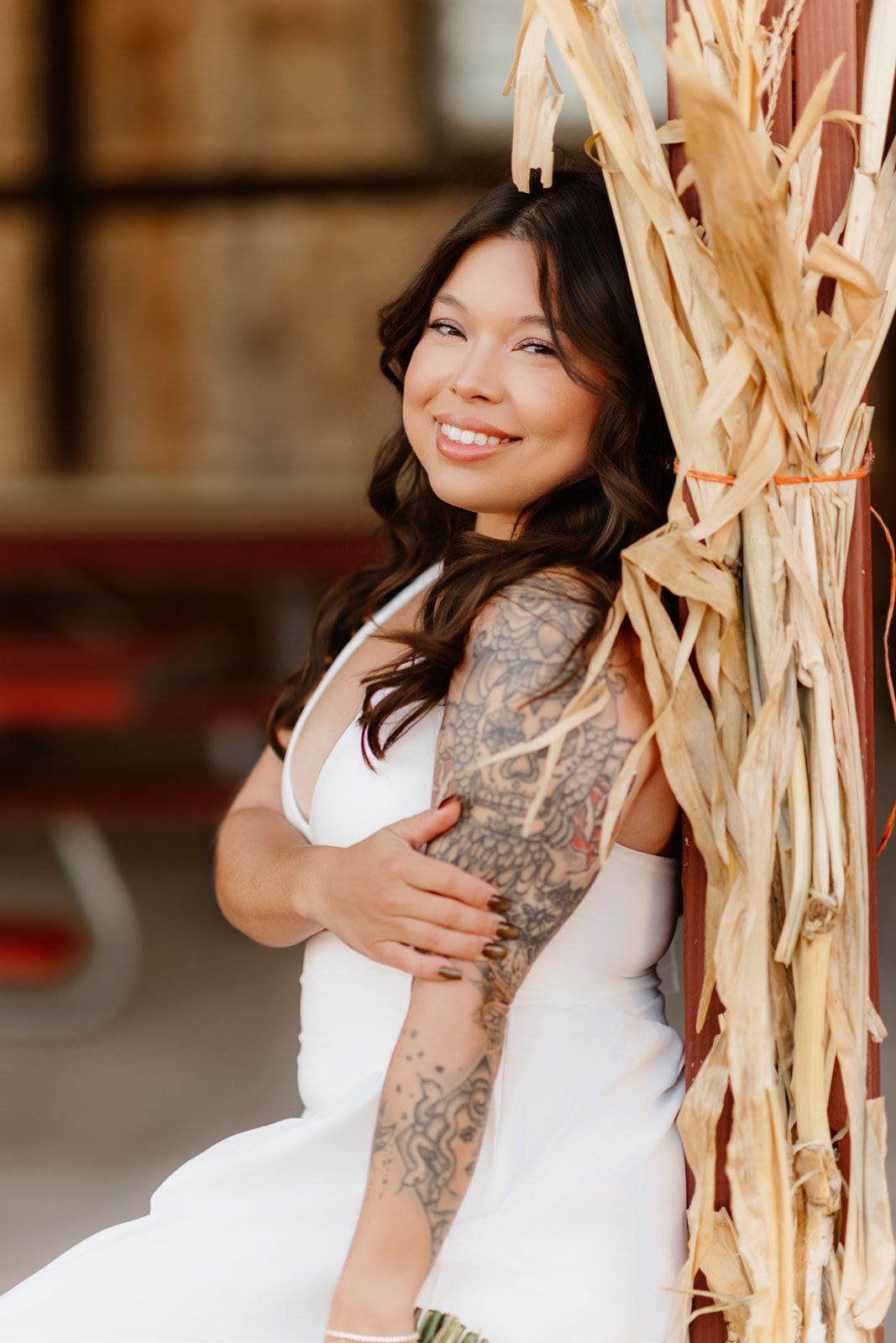 Smiling bride in white dress with tattoo sleeve leans against rustic corn stalk decoration at barn wedding venue.