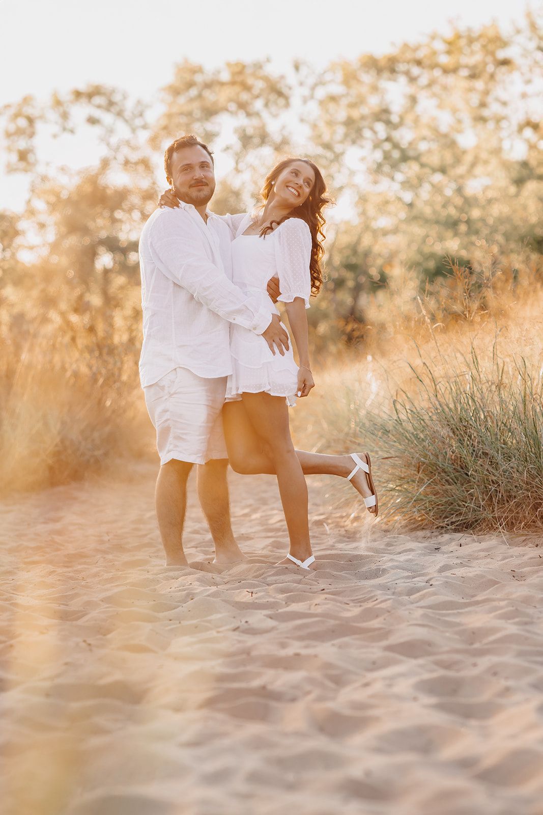 Happy couple in white outfits embracing on a sandy beach path at golden hour sunset.