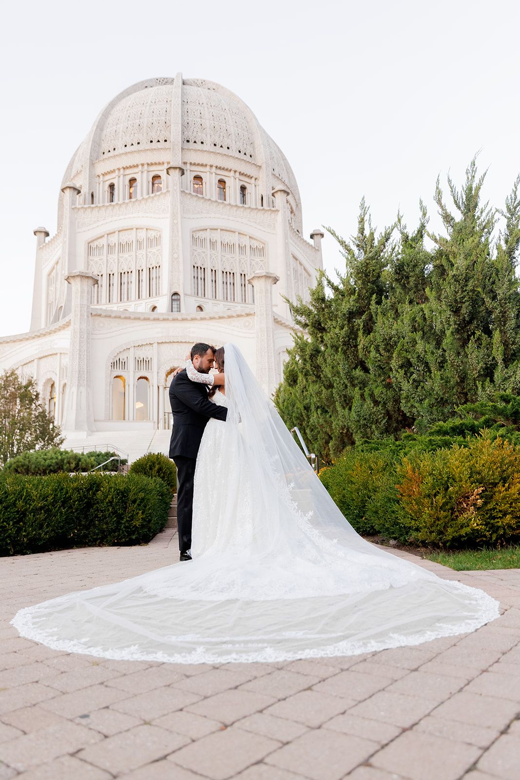 Bride and groom embrace in front of the white domed Bahai Temple, with long flowing veil trailing on stone pavers.