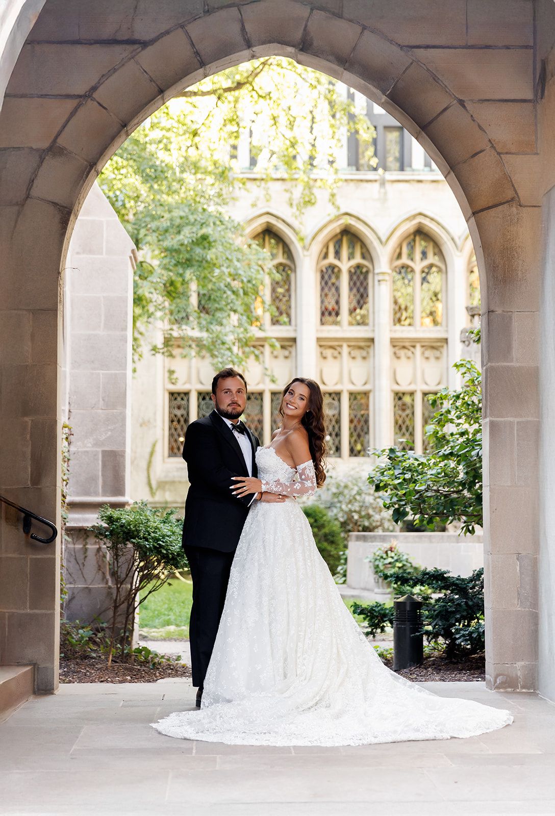 Bride and groom pose under Gothic stone arch in wedding attire, lush courtyard and ornate windows in background.
