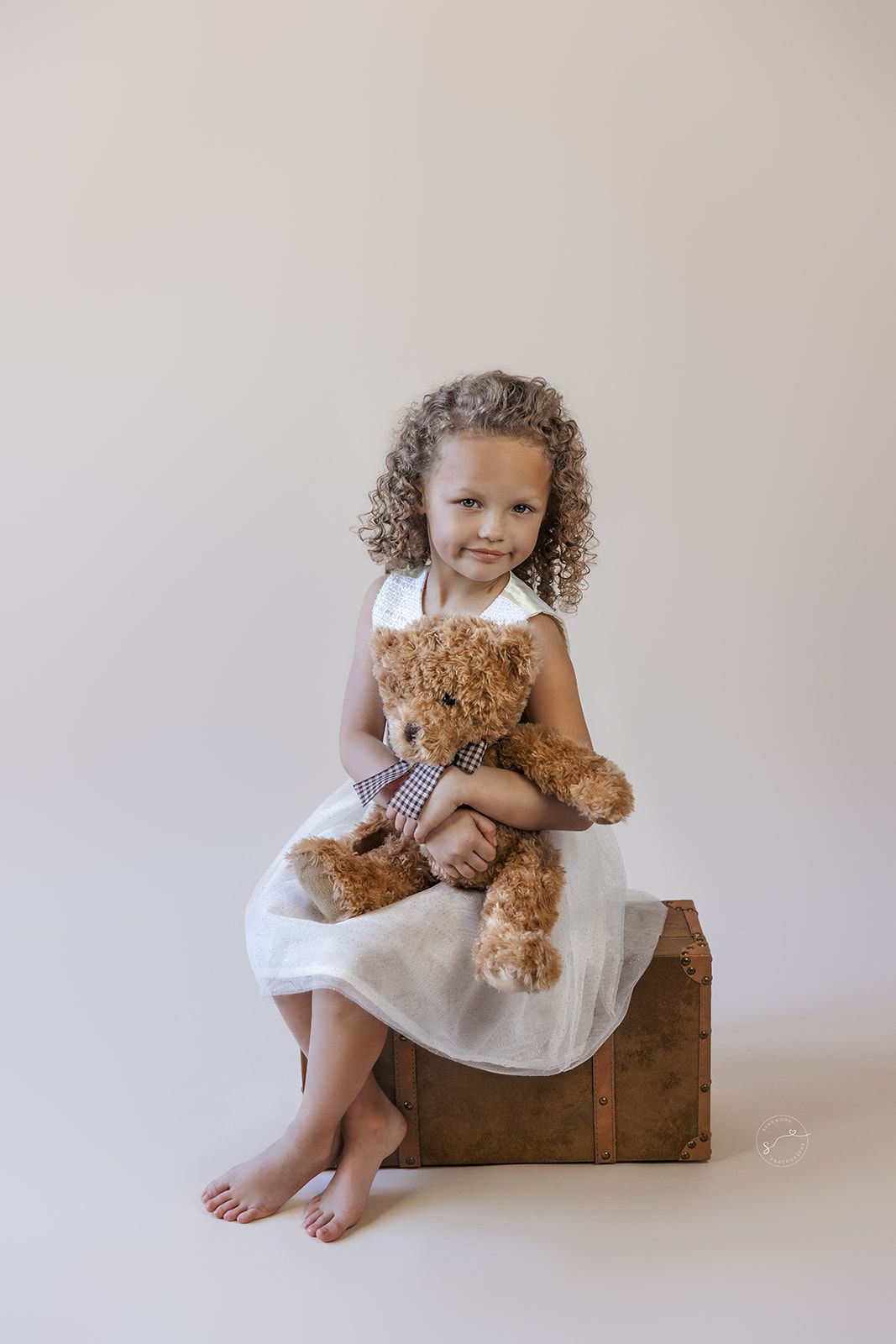 Young girl in white dress hugging brown teddy bear, sitting on wooden crate with curly hair, barefoot on white background.