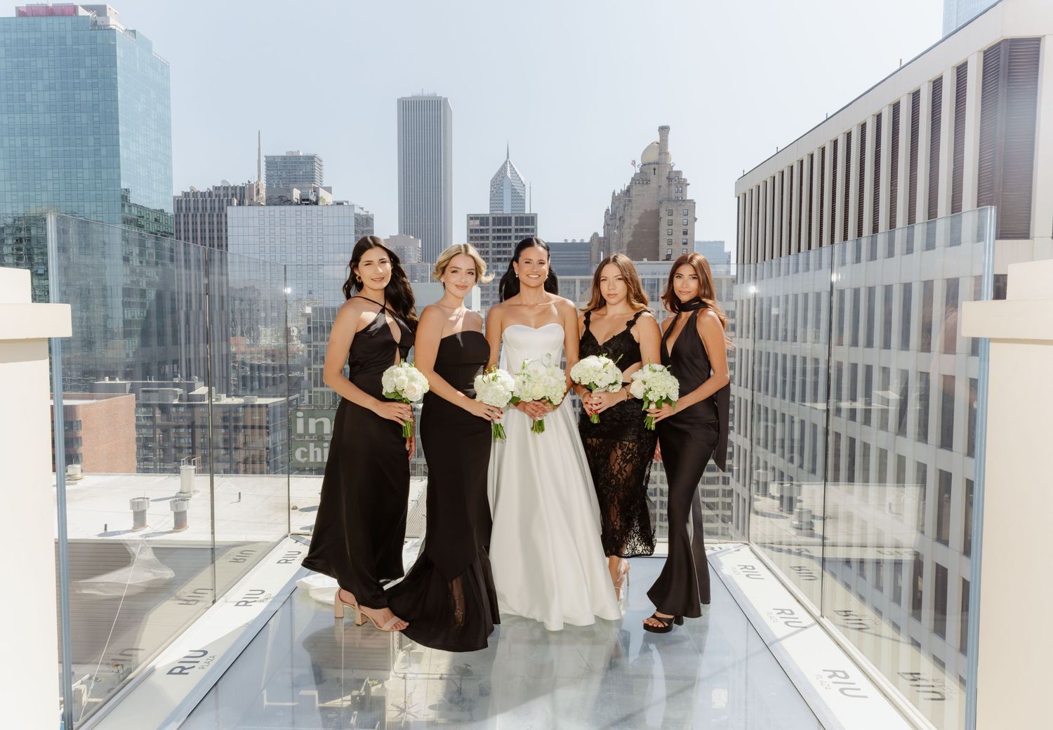 Bride and four bridesmaids in black dresses walking on glass rooftop with Chicago skyline.