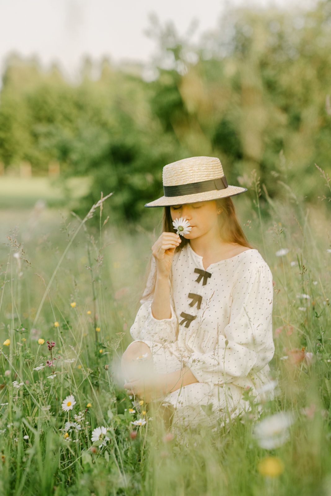 Young woman in straw hat and white dress smelling a daisy while sitting in a green meadow.