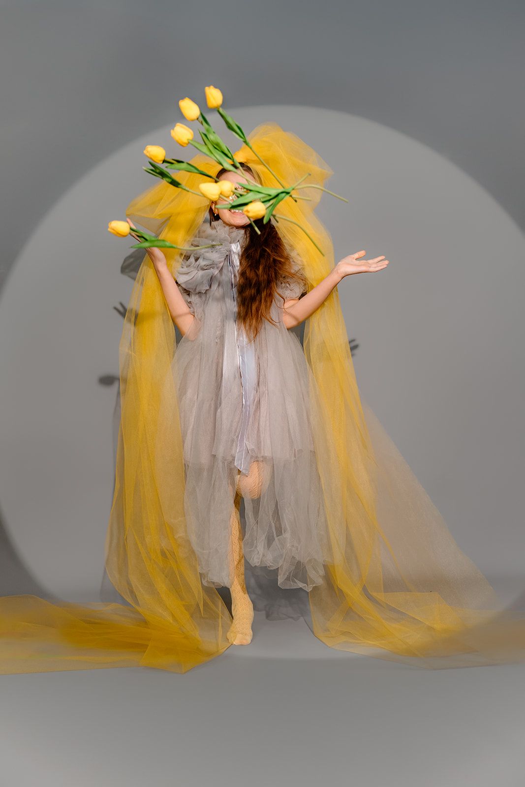 Woman in sheer gray dress with yellow tulle and tulips covering her face poses gracefully in studio setting.