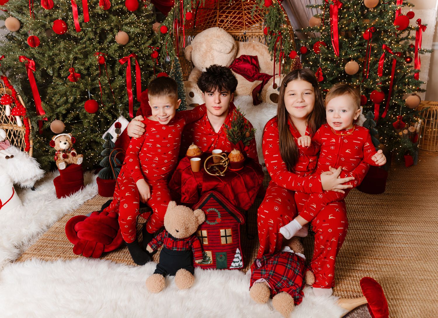 Four children in matching red Christmas pajamas pose together in front of a decorated holiday tree with gifts.