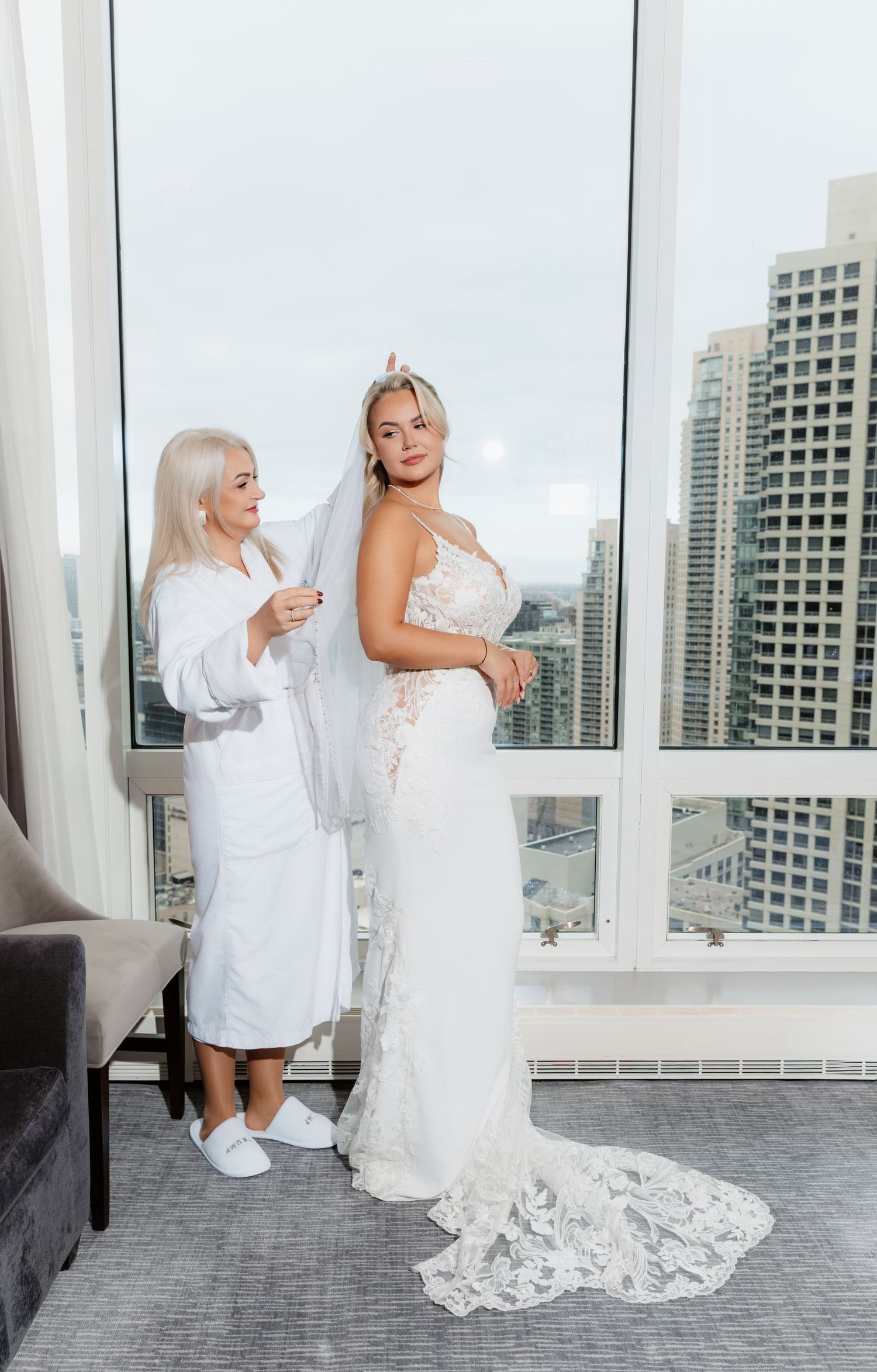 Older woman in white robe adjusting bride's veil in elegant lace wedding gown by city window.