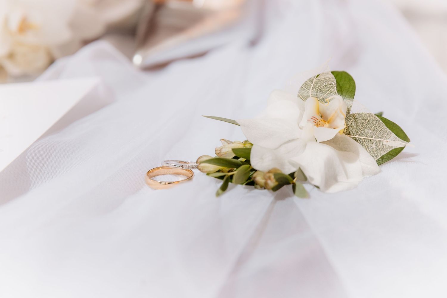 Two gold wedding bands rest on a white orchid boutonniere atop a soft bridal veil.