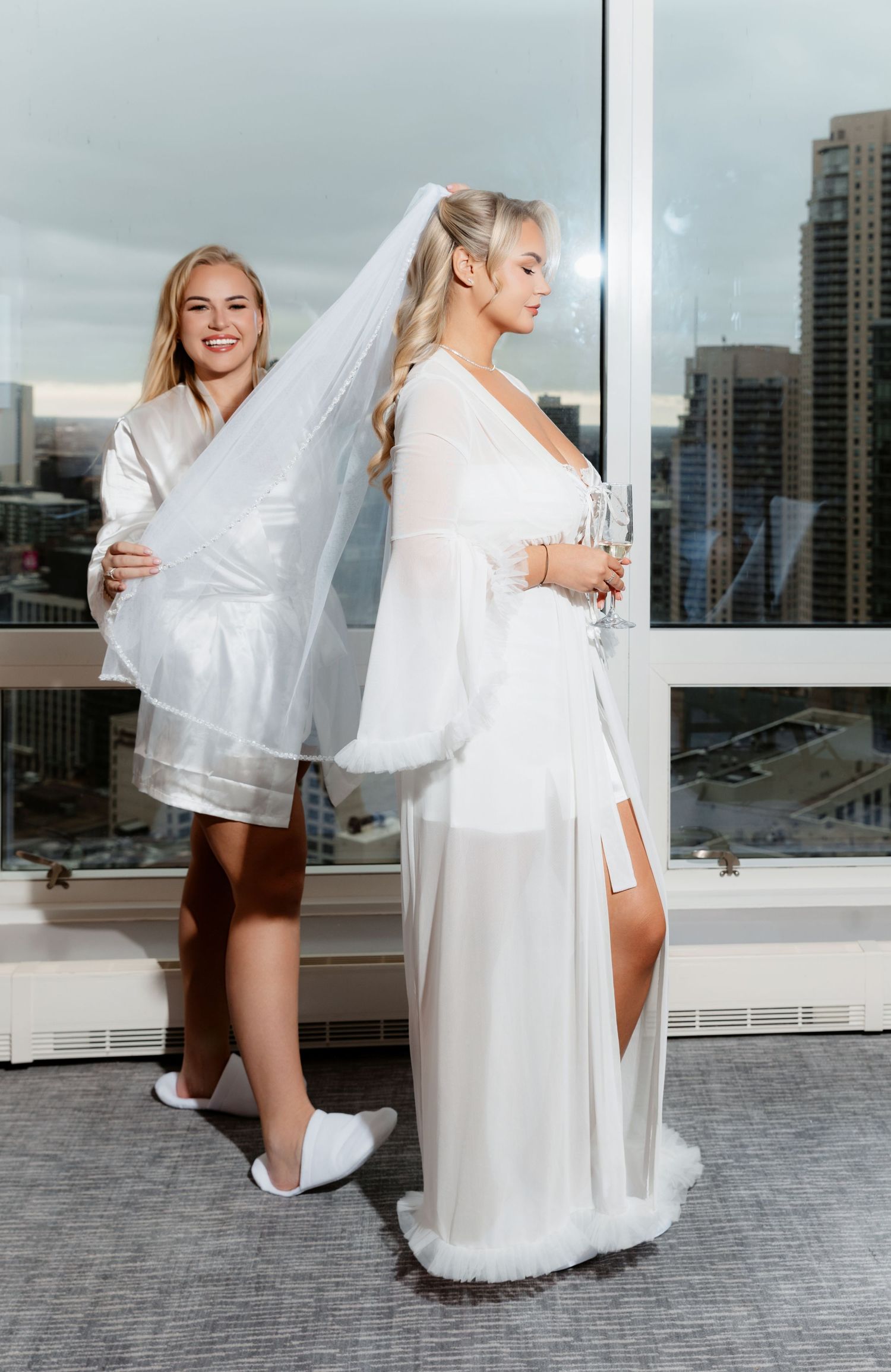 Bridesmaid adjusting veil on bride in white sheer robe by floor-to-ceiling hotel window.