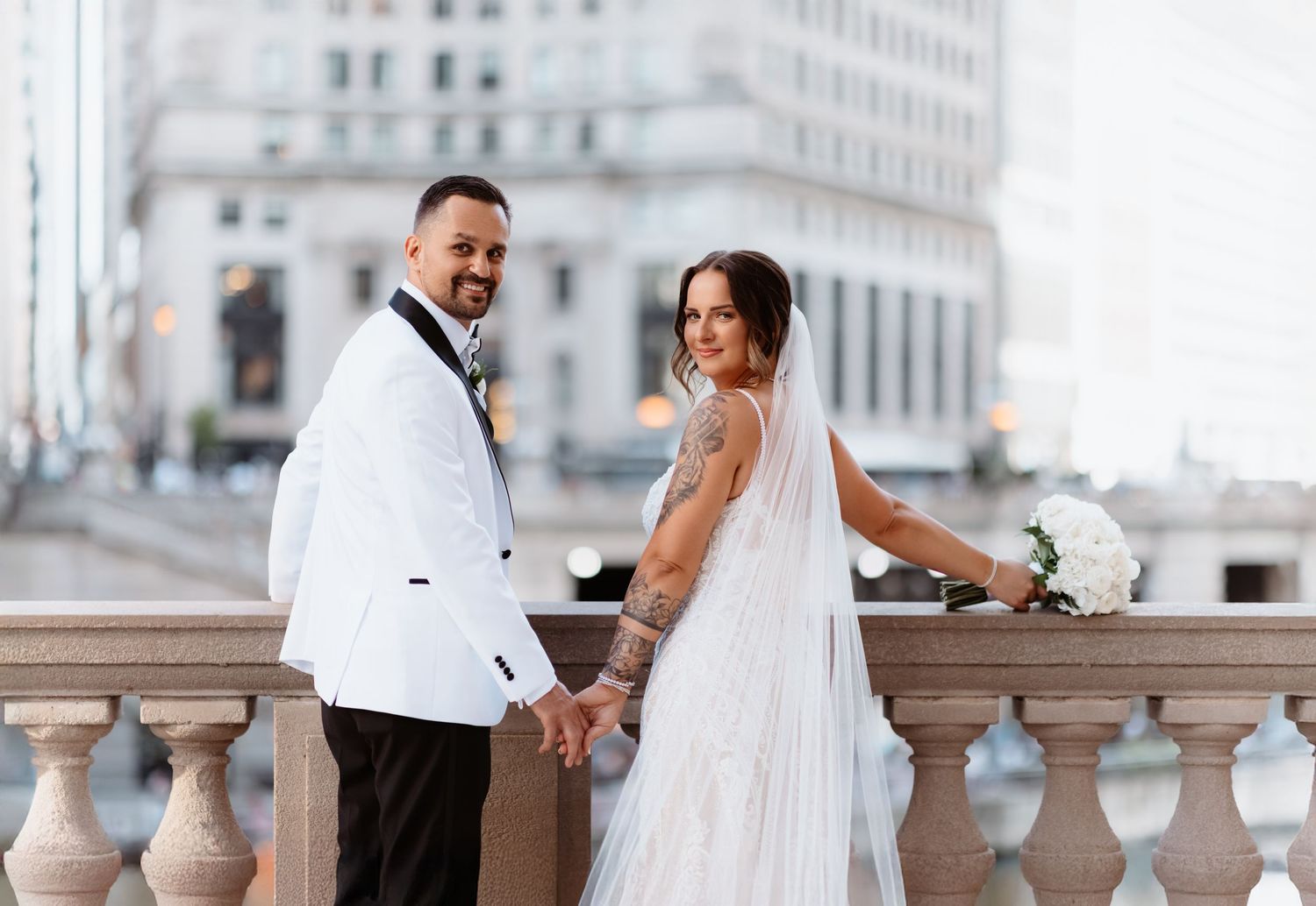 Bride and groom holding hands smiling on urban bridge with city buildings behind.