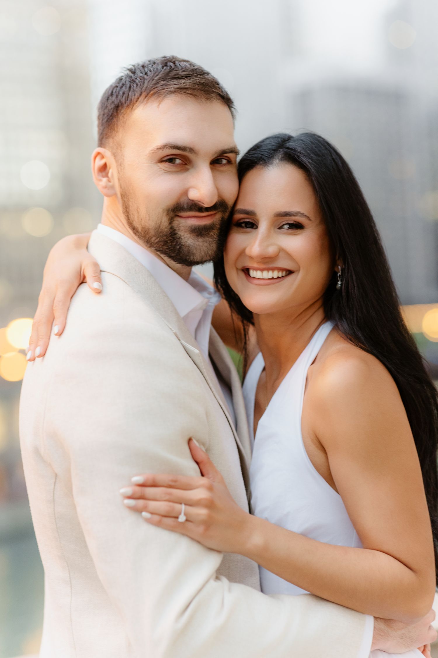Woman in white dress hugging man in cream suit, both smiling at camera with city backdrop.