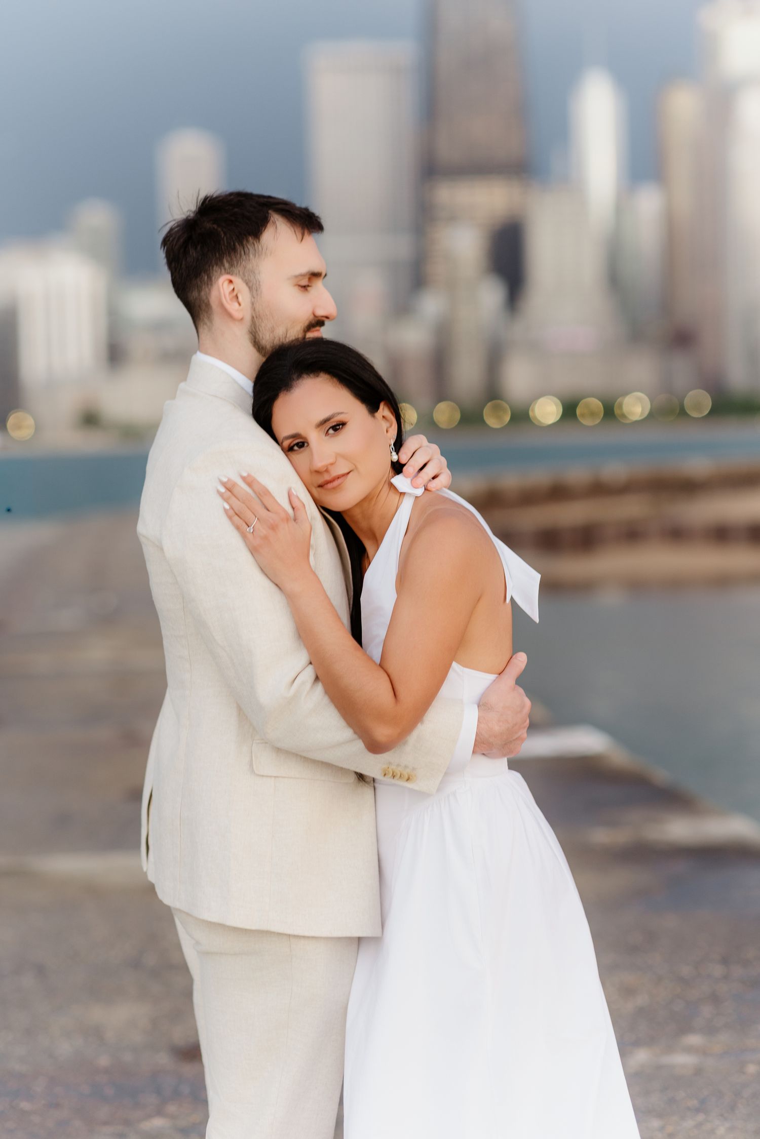 Woman in white dress rests head on mans chest as they embrace with blurred Chicago skyline behind.