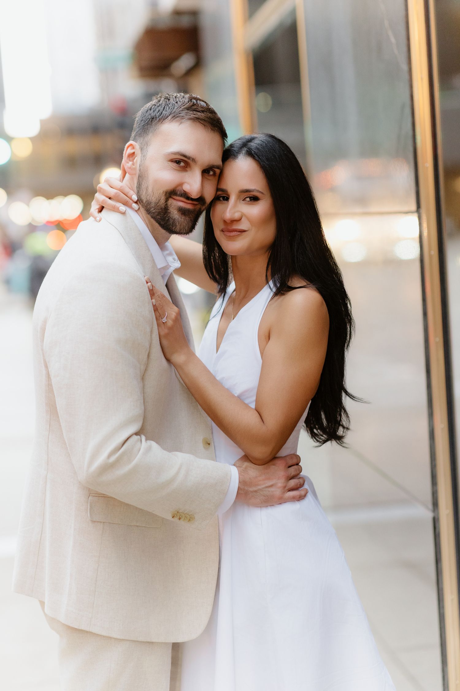 Stylish engaged couple embracing and smiling beside a gold-framed building on a city sidewalk.