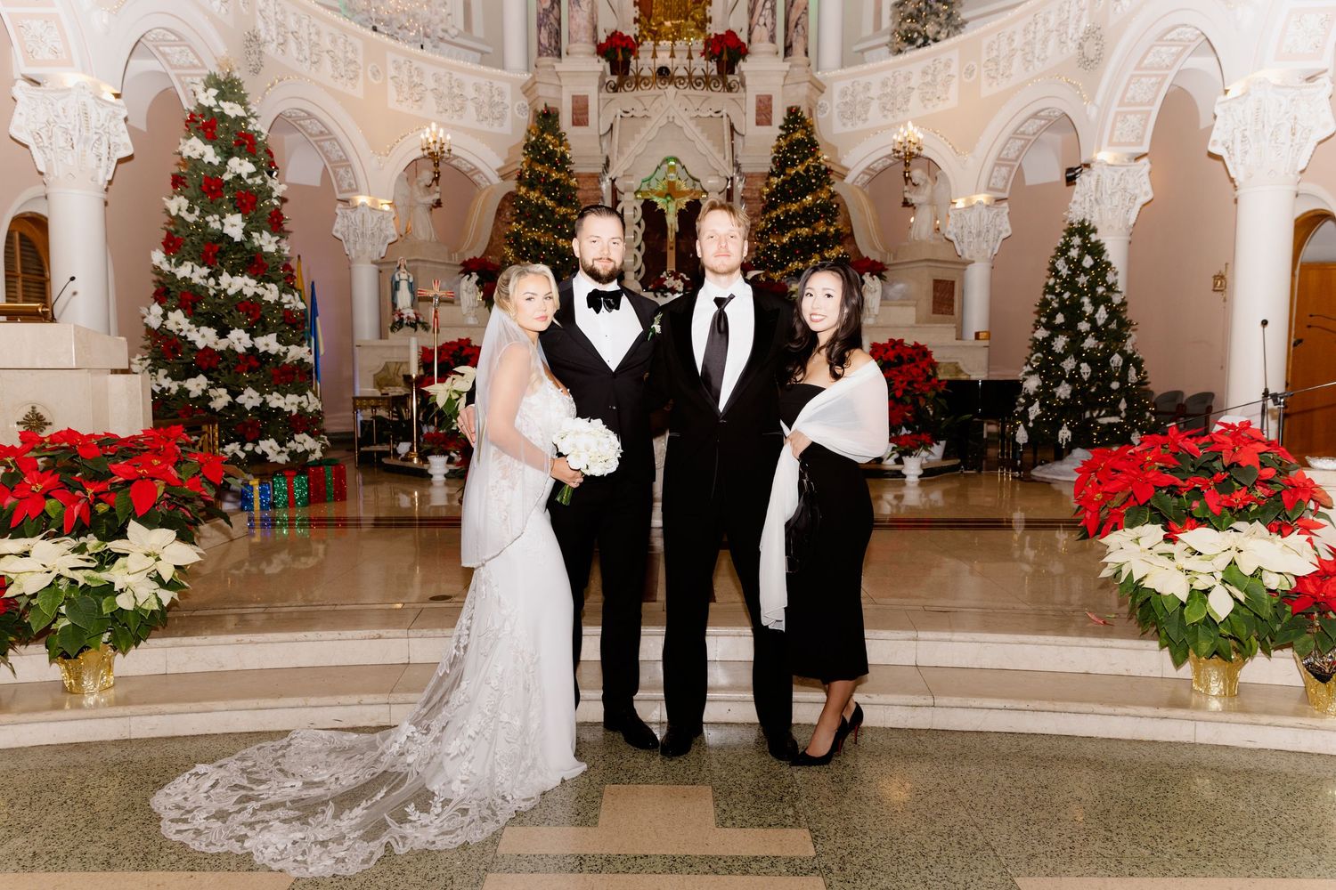 Wedding party of four posing inside Christmas-decorated church with poinsettias and holiday trees.