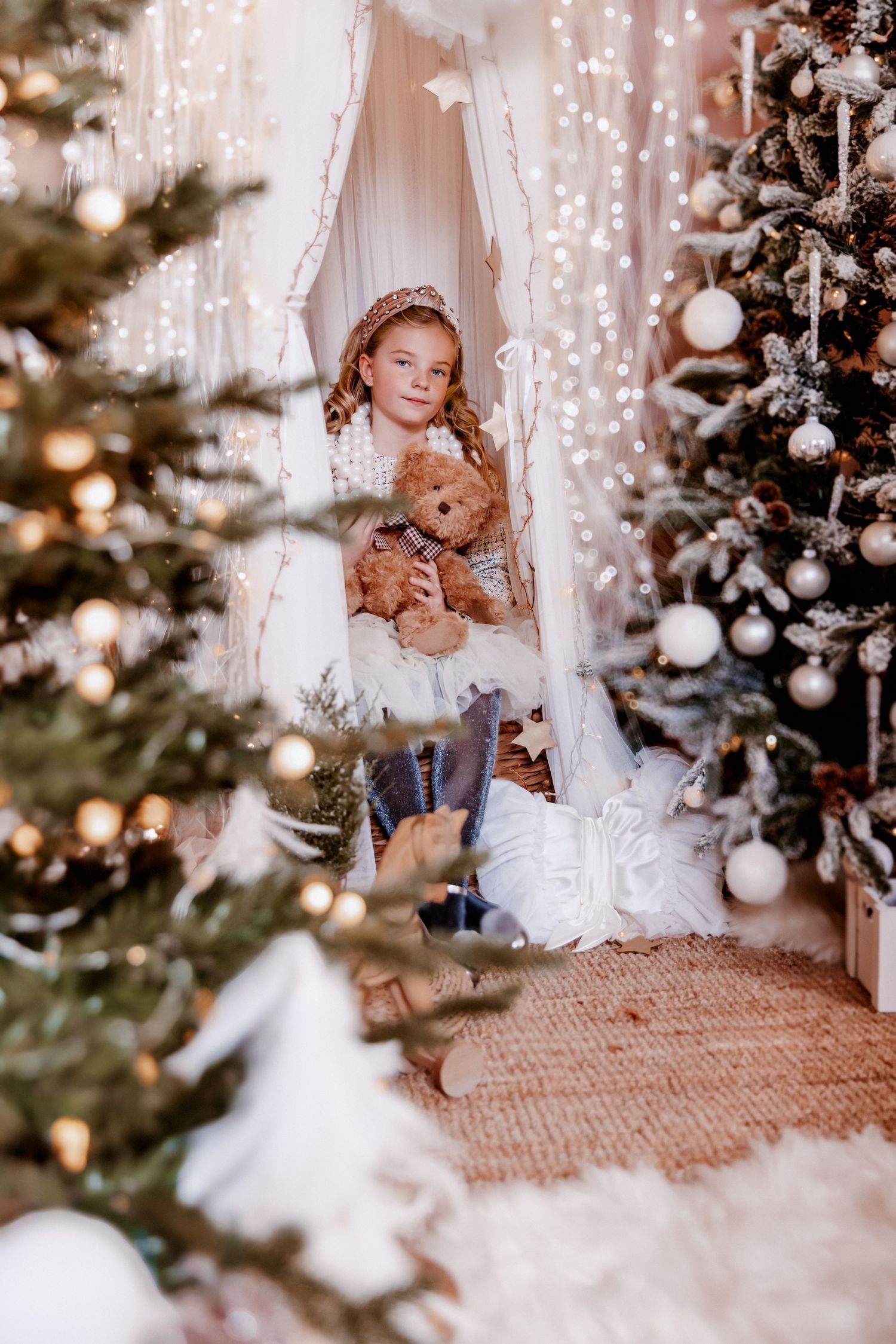 Curly-haired girl hugs teddy bear inside canopy surrounded by glowing Christmas trees.
