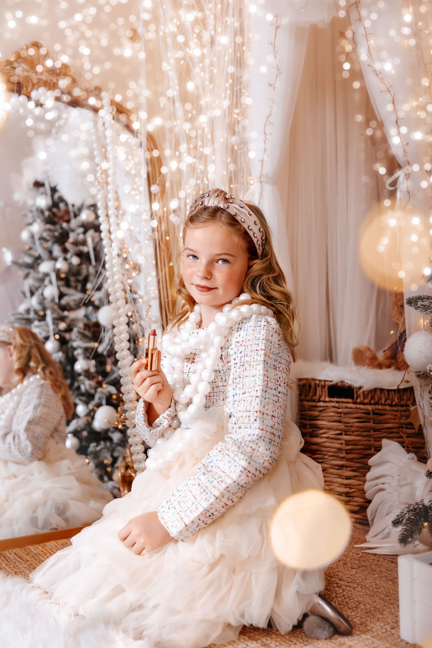 Blonde girl in white tweed jacket holds nutcracker amid sparkling Christmas light backdrop.
