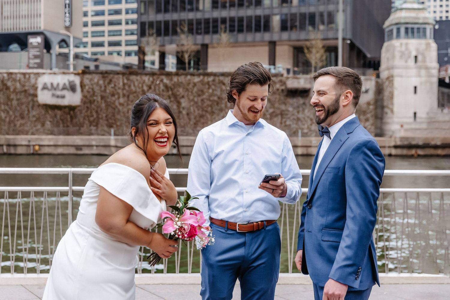 Bride in white dress laughs with two men in blue suits during an outdoor urban wedding ceremony.
