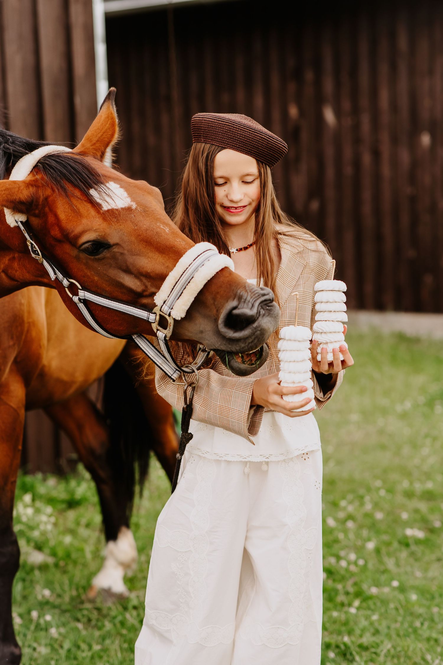 Girl in brown beret offers stacked marshmallow treats to a chestnut horse beside a barn.