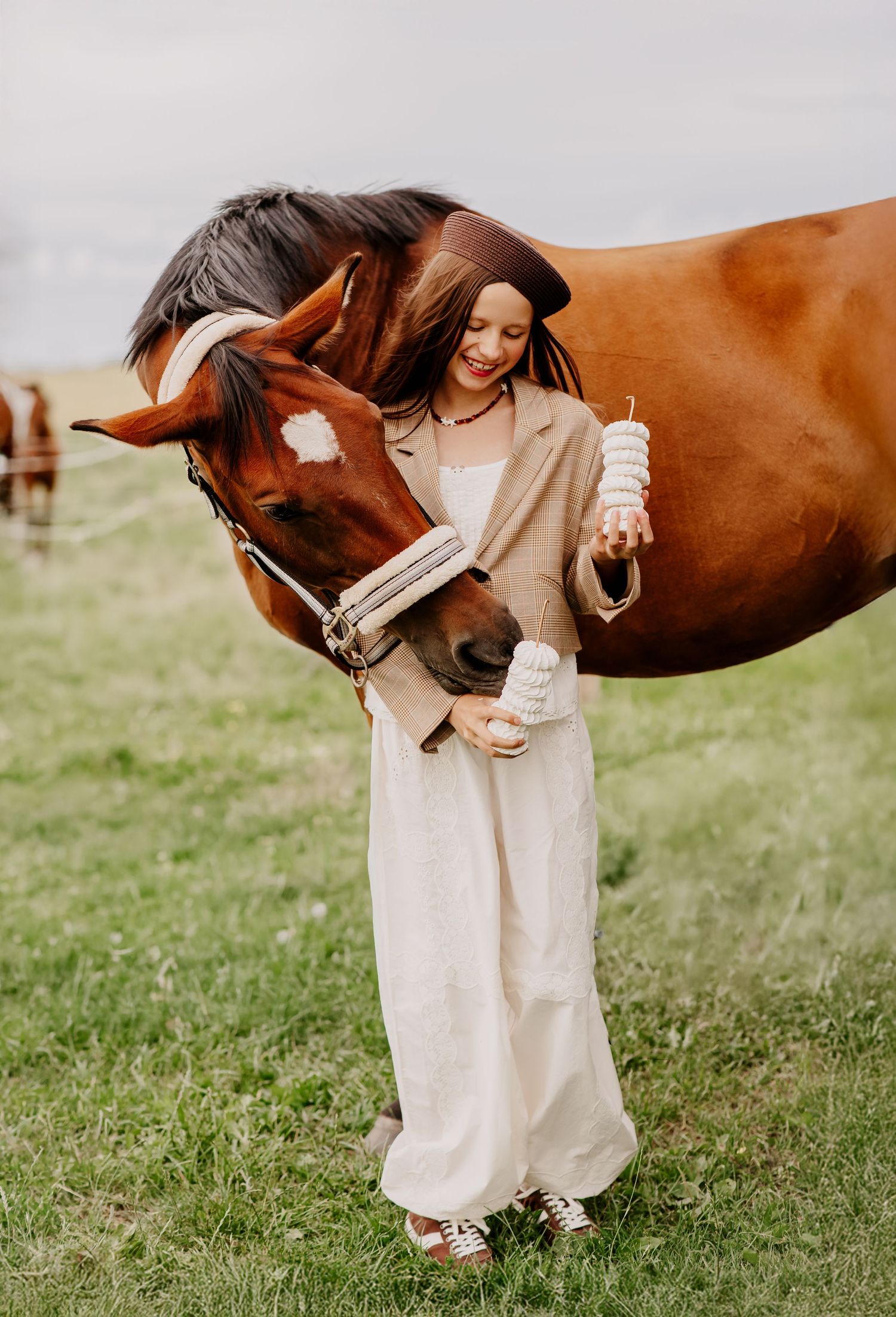 Young woman in a beige outfit feeding a chestnut horse in a green meadow.