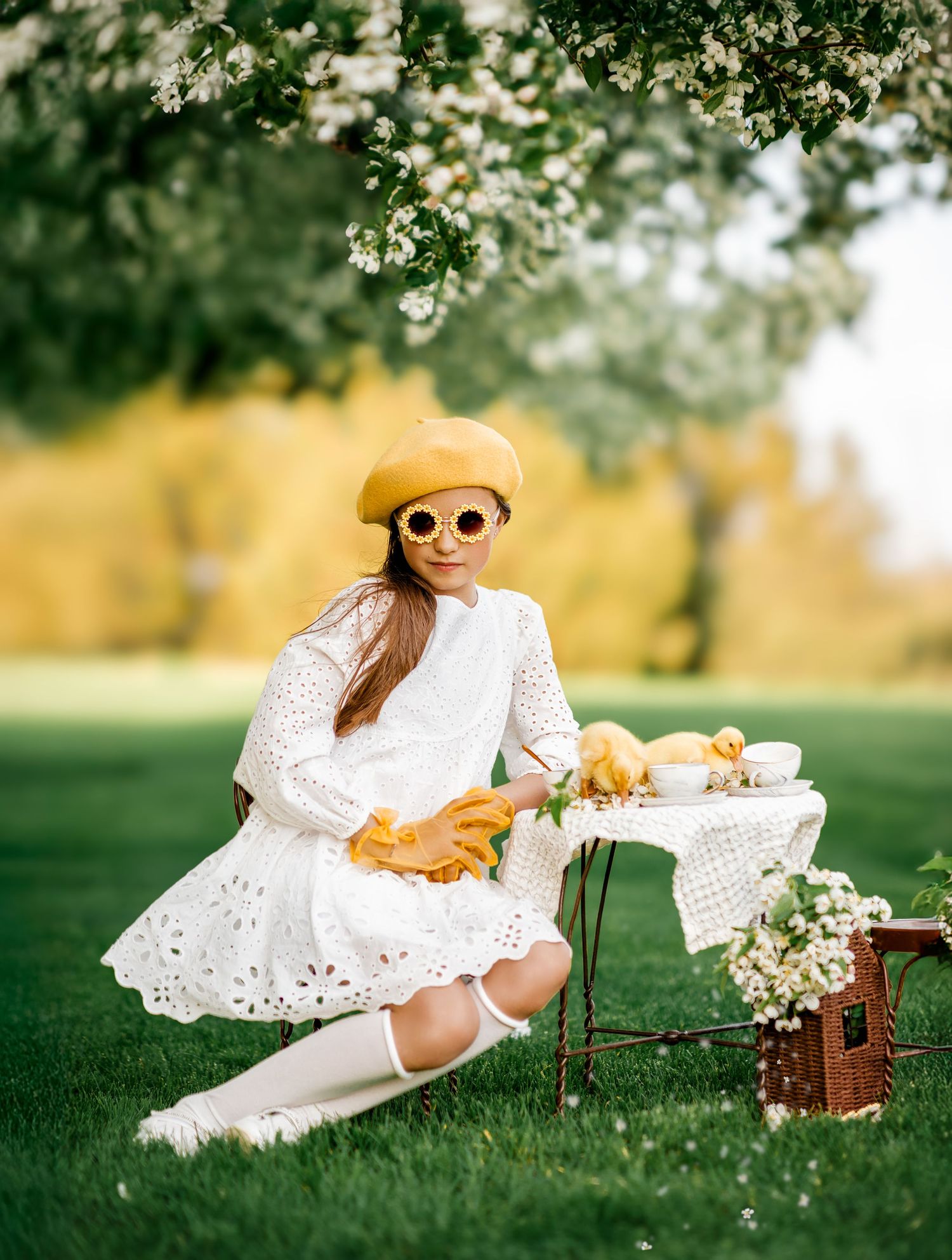 Girl in white dress, yellow beret and sunglasses sits on grass at picnic table with ducklings under blooming tree.