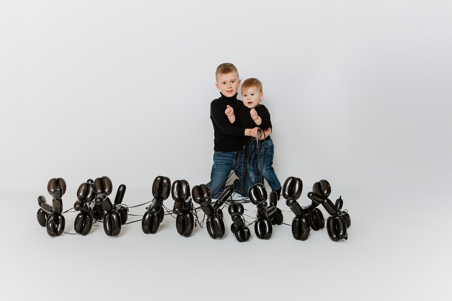 Two young boys in black sweaters surrounded by black balloon animal dogs on white background.