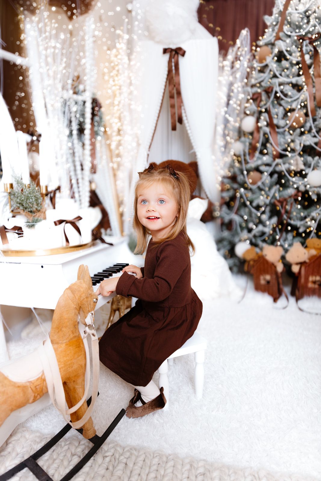 Young girl in brown dress plays toy piano beside decorated Christmas tree with festive lights and snowy decor.