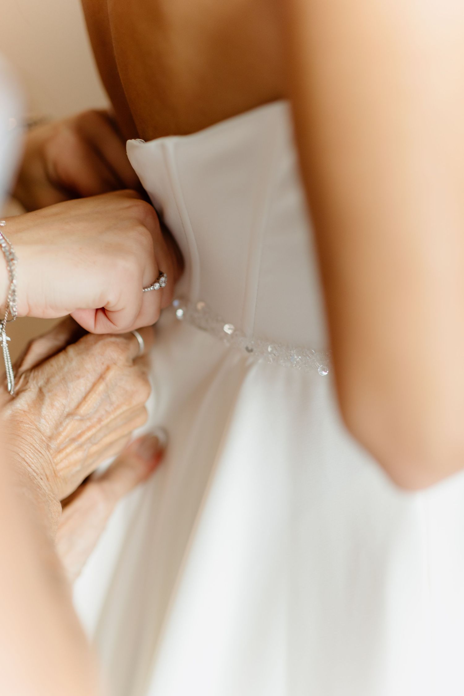 Elderly hands fastening pearl buttons on the back of a white bridal gown during wedding preparations.