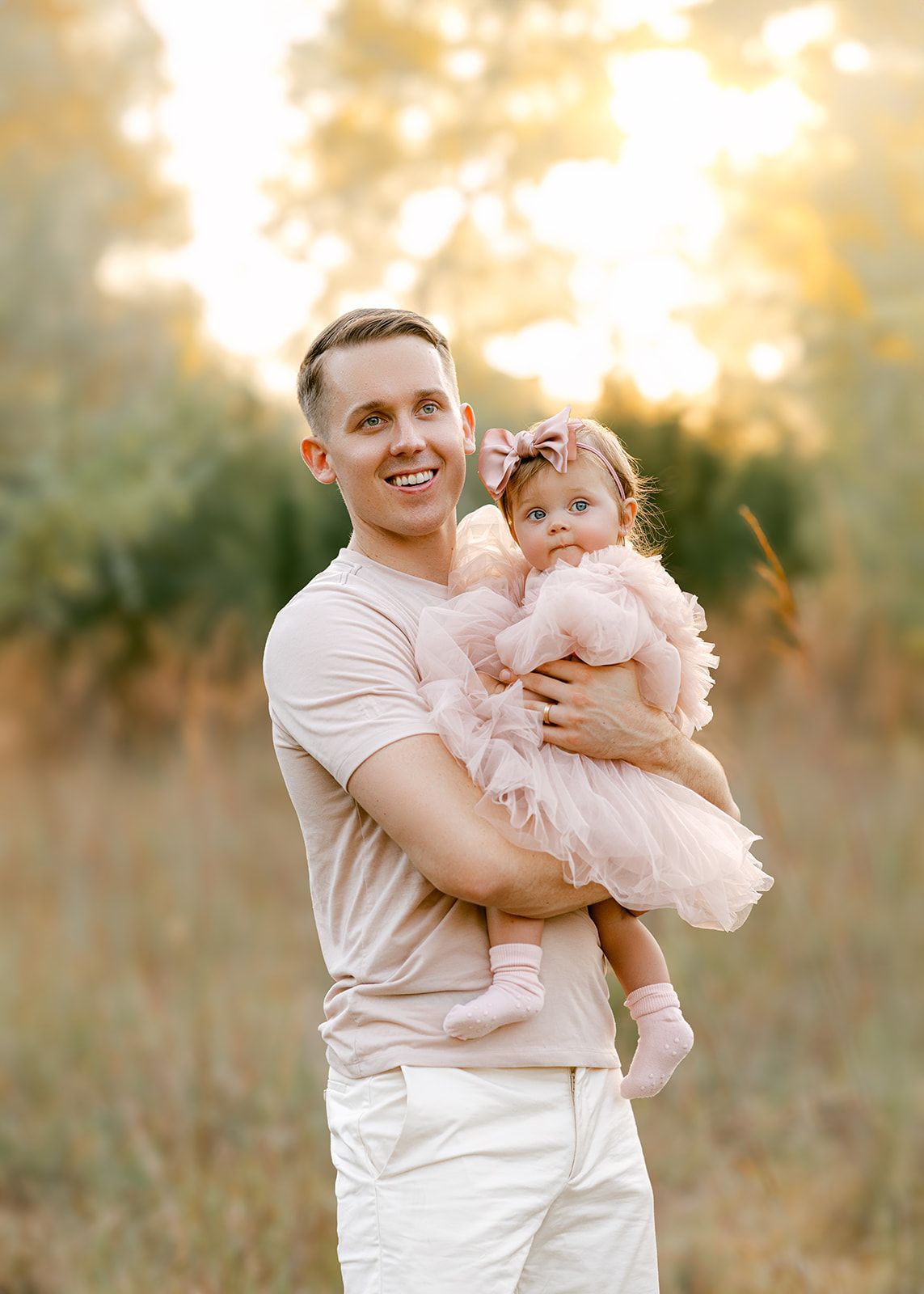 Happy father in beige shirt holds baby girl in pink tulle dress in golden sunlit field.