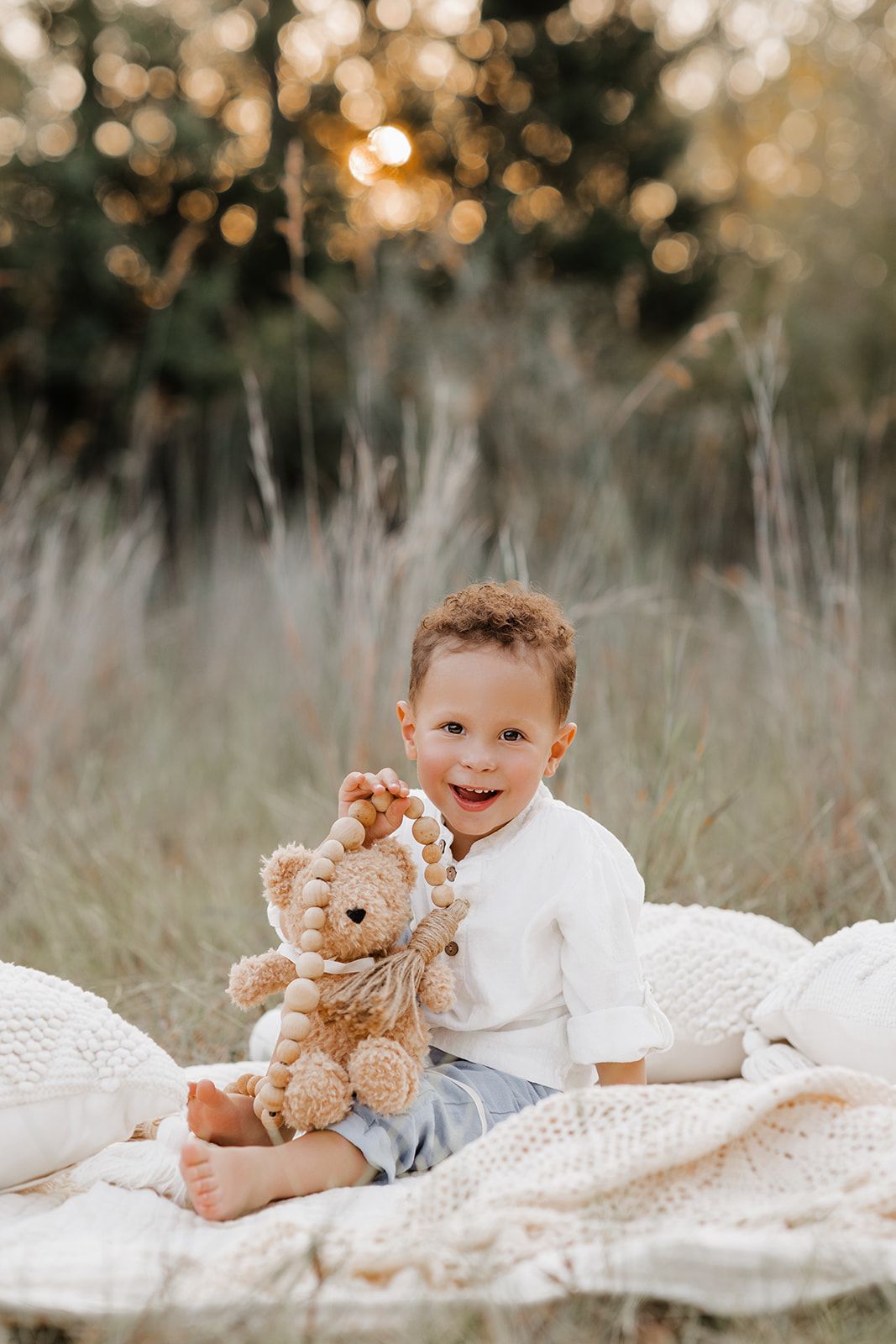 Smiling toddler boy sitting on white blanket in field holding teddy bear at golden hour sunset.