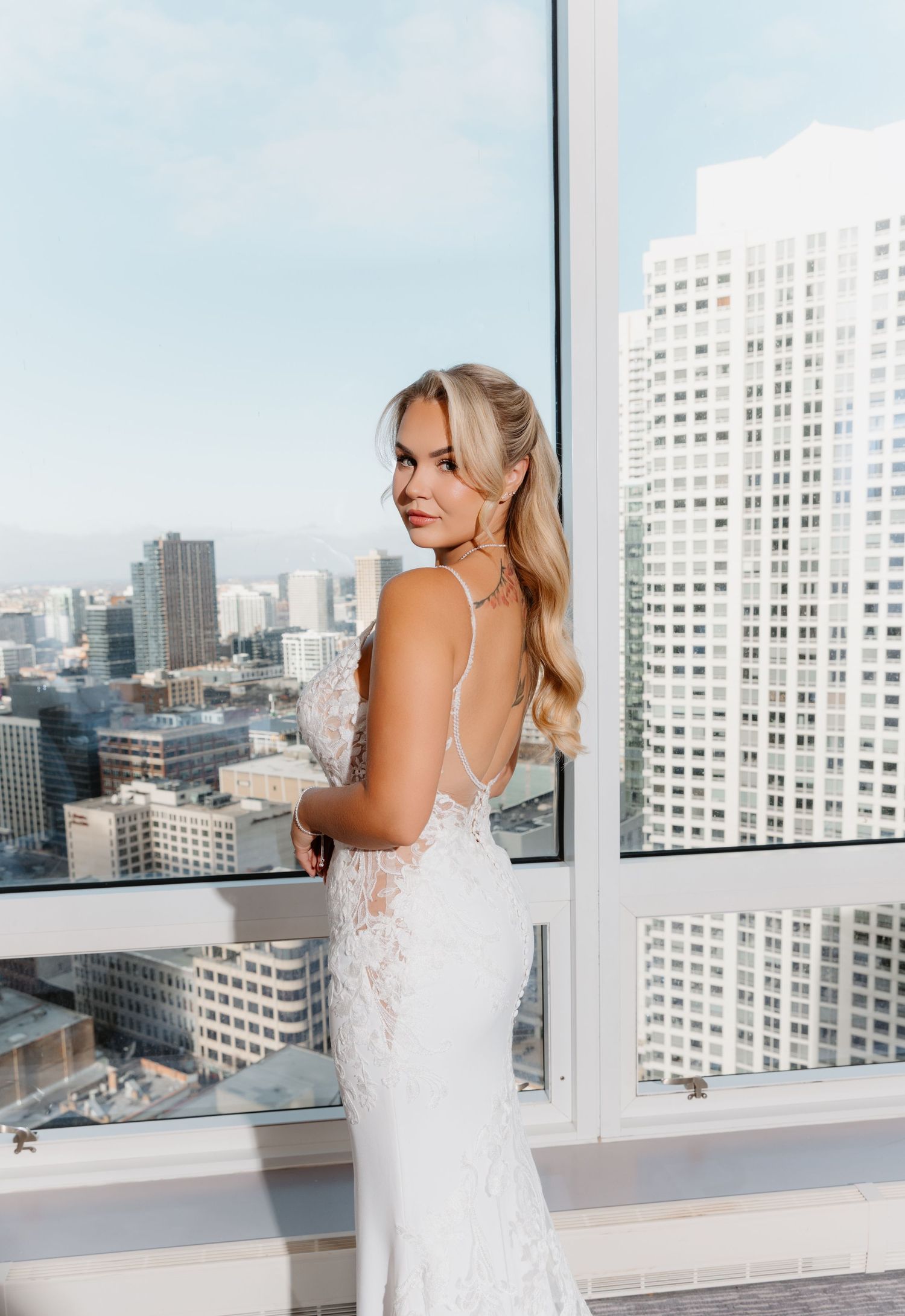 Blonde bride in backless lace wedding gown posing by floor-to-ceiling windows with city skyline.