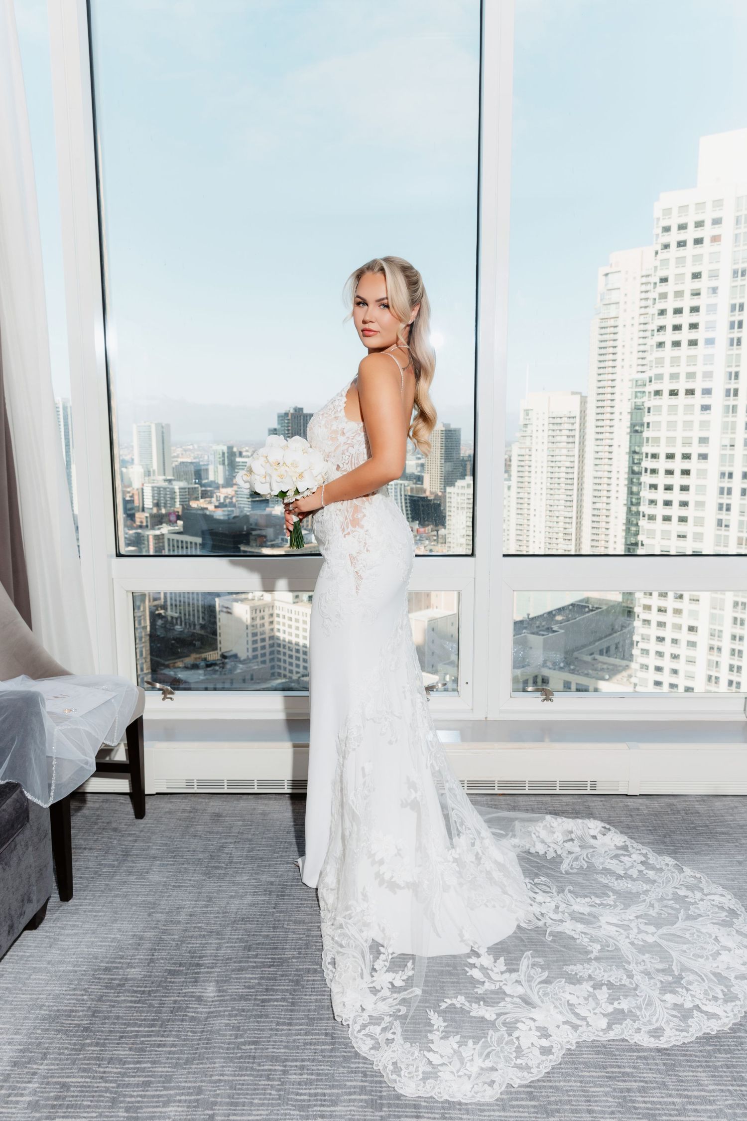 Blonde bride in sleeveless lace wedding gown holding white bouquet poses before high-rise city view.