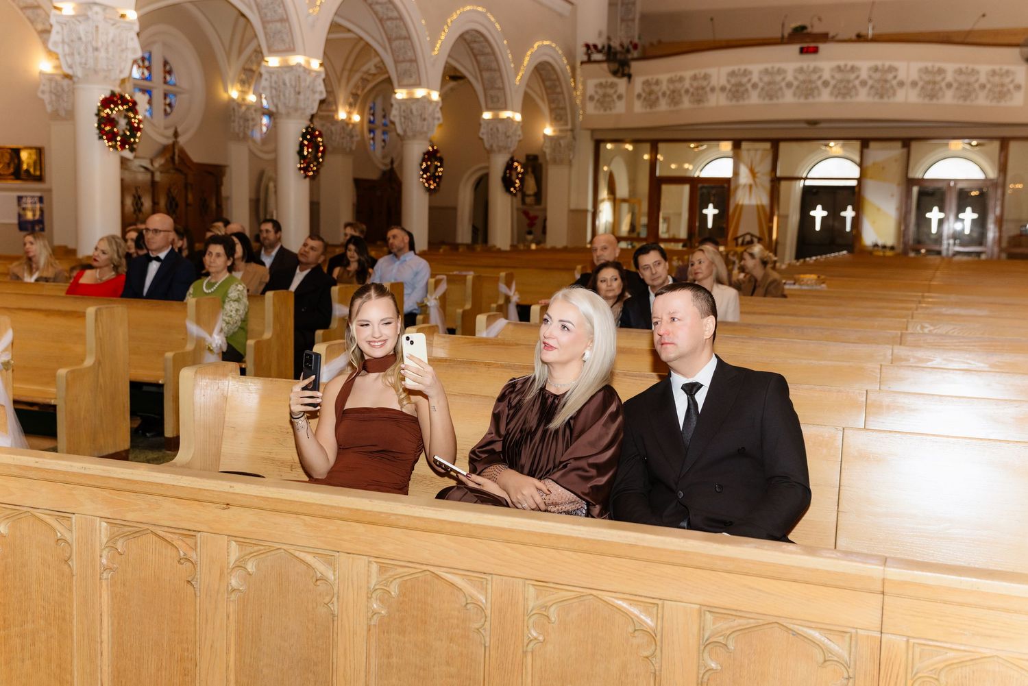 Wedding guests seated in ornate church pews capturing ceremony moments on phones.