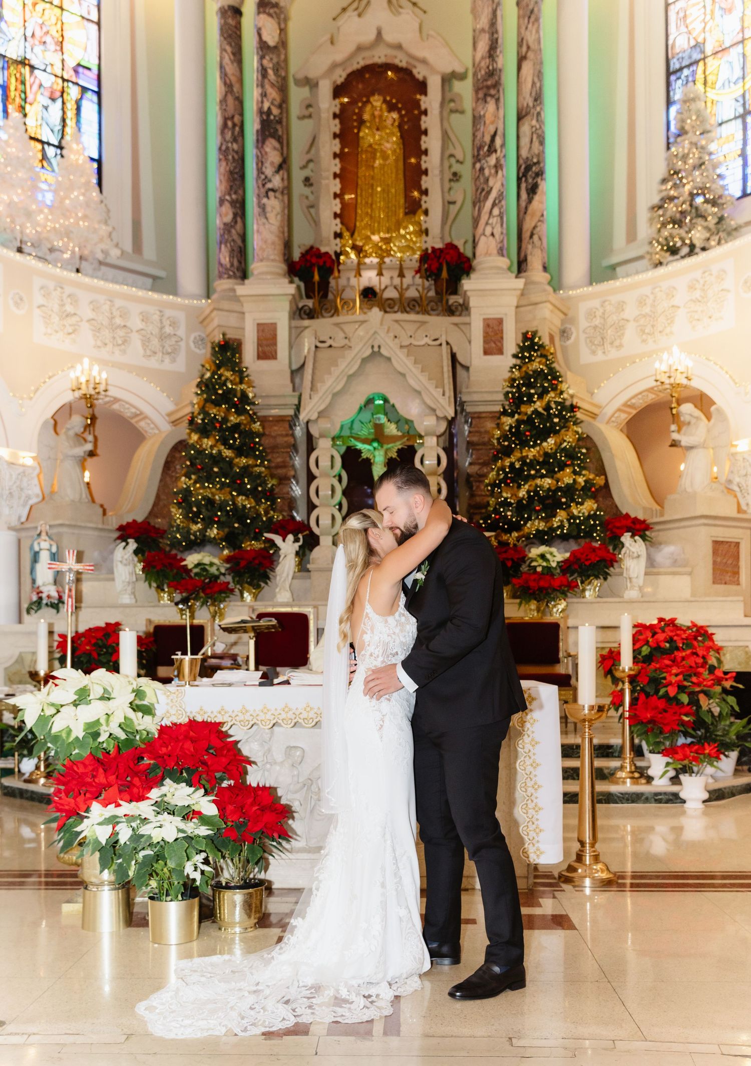 Bride and groom kissing at ornate church altar decorated with Christmas trees and poinsettias.