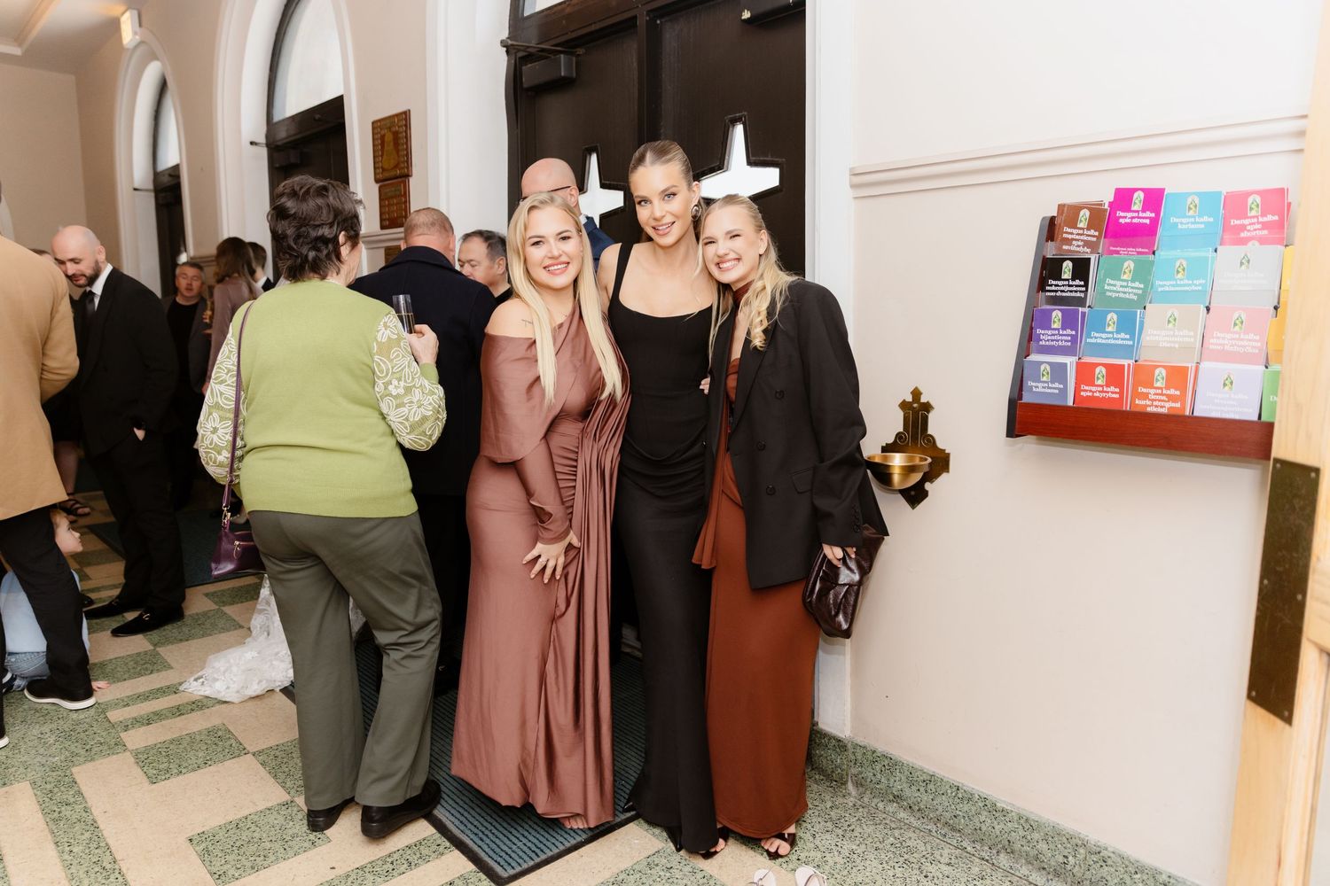 Three stylish women posing together at formal event near colorful display.