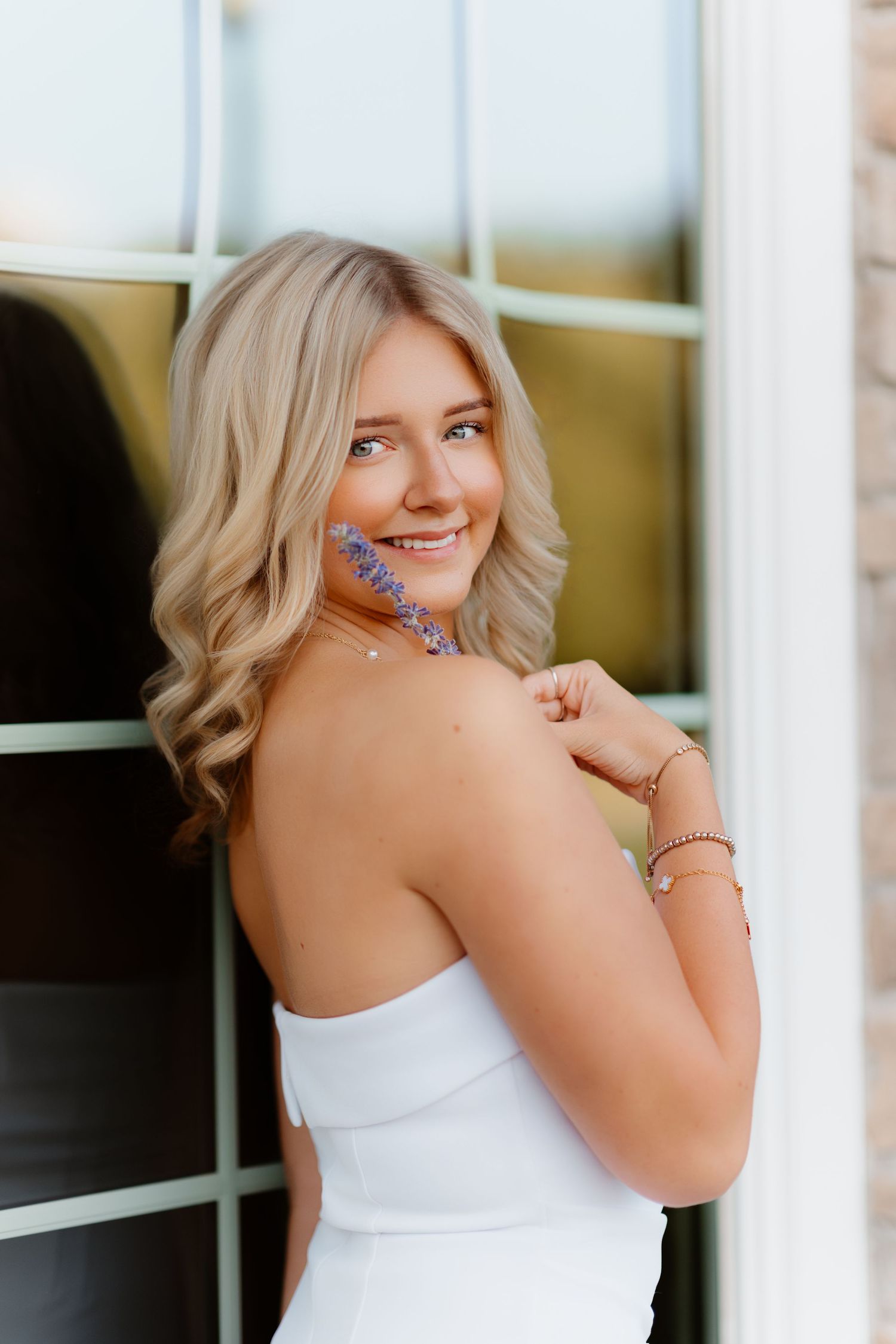 Blonde woman in white strapless dress smiling over shoulder holding purple flowers.